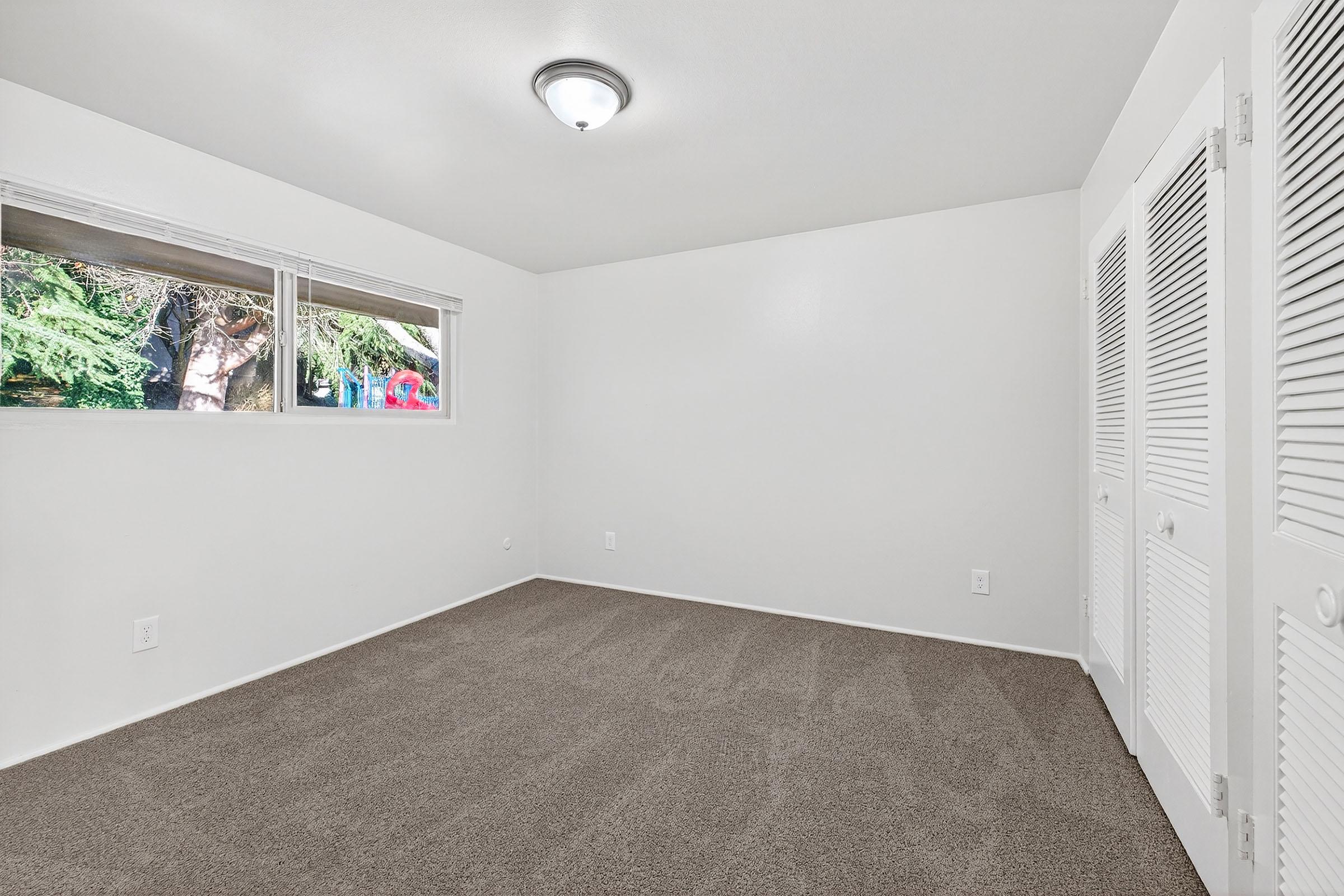 Empty room with light gray carpet and white walls. Two windows allow natural light, revealing greenery outside. A ceiling light fixture is visible. There are two sets of white closet doors on one wall, and the overall space is clean and uncluttered, ready for furnishings.