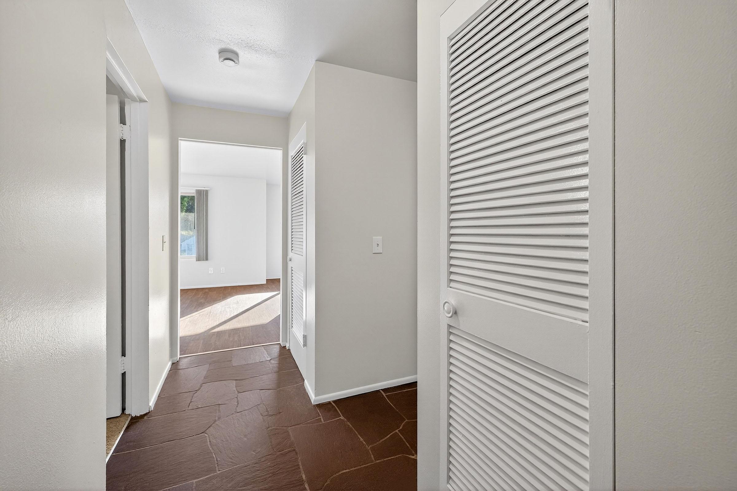 A hallway featuring light-colored walls, a tan stone floor, and several doors, including a closet with white louvered doors. At the end of the hallway, a doorway leads to a room with larger windows, indicating natural light. The overall atmosphere is bright and inviting.