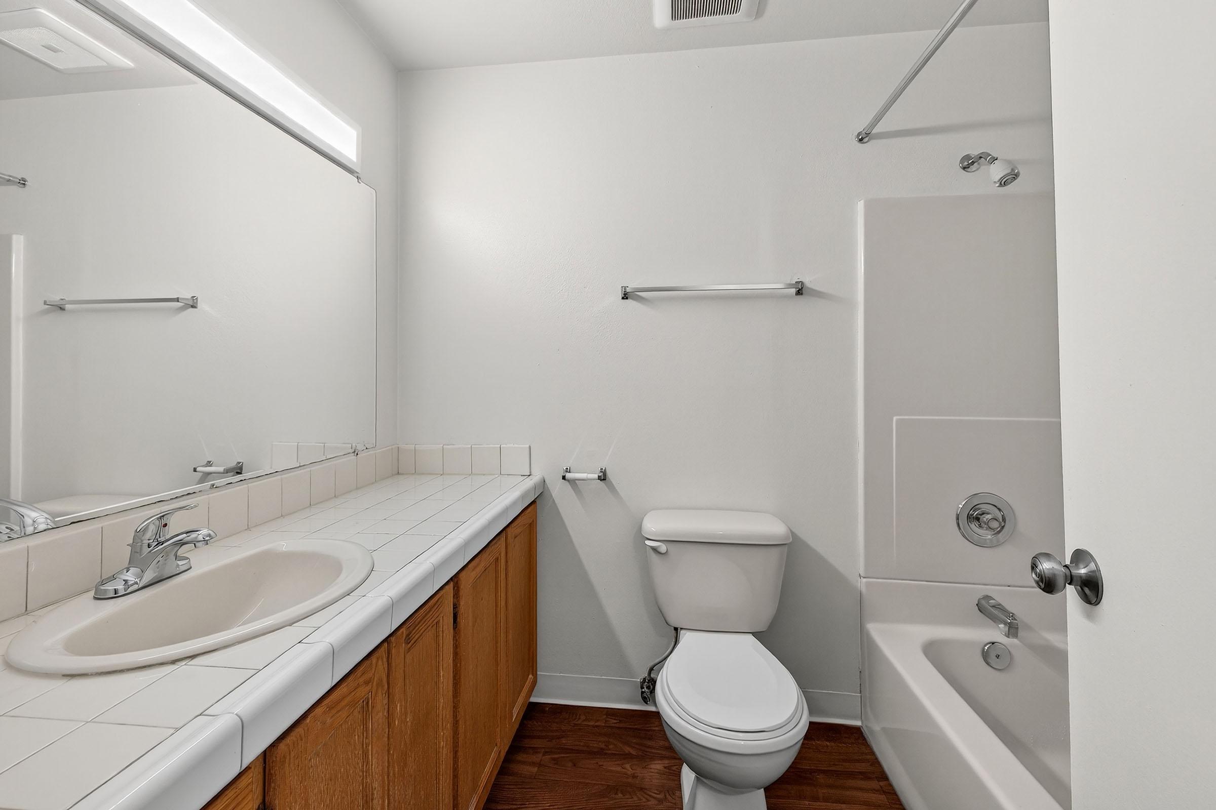A clean, modern bathroom featuring a white tiled countertop with a sink and mirror, a toilet, a bathtub, and a shower. The walls are painted light colors, and wooden flooring adds warmth to the space. Simple fixtures and minimalistic design create a spacious feel.