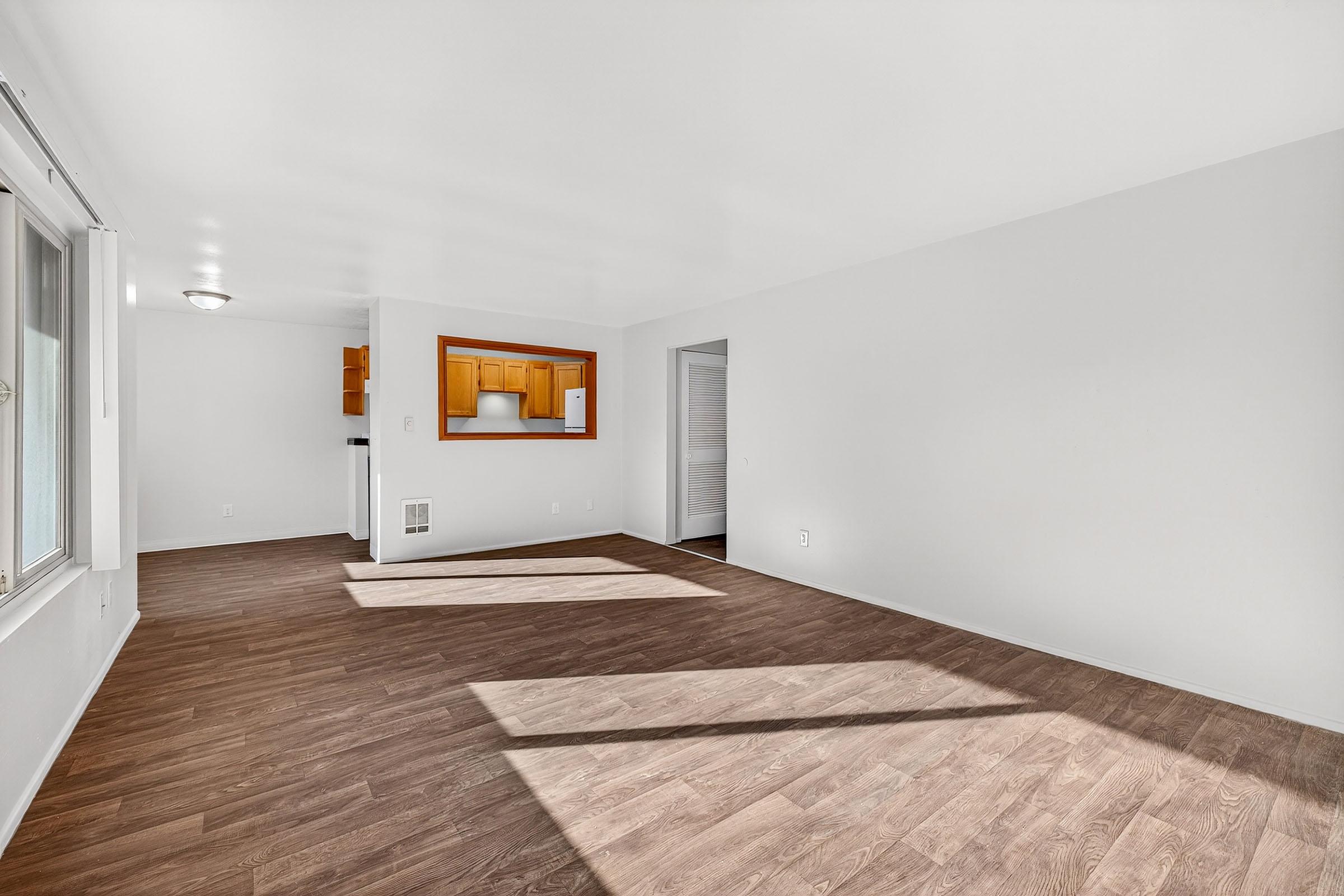 A spacious, empty room with white walls and a brown laminate floor. Natural light streams in through a window, casting shadows on the floor. In the background, there is an opening leading to a kitchen area with wooden cabinets. The overall atmosphere is bright and inviting.