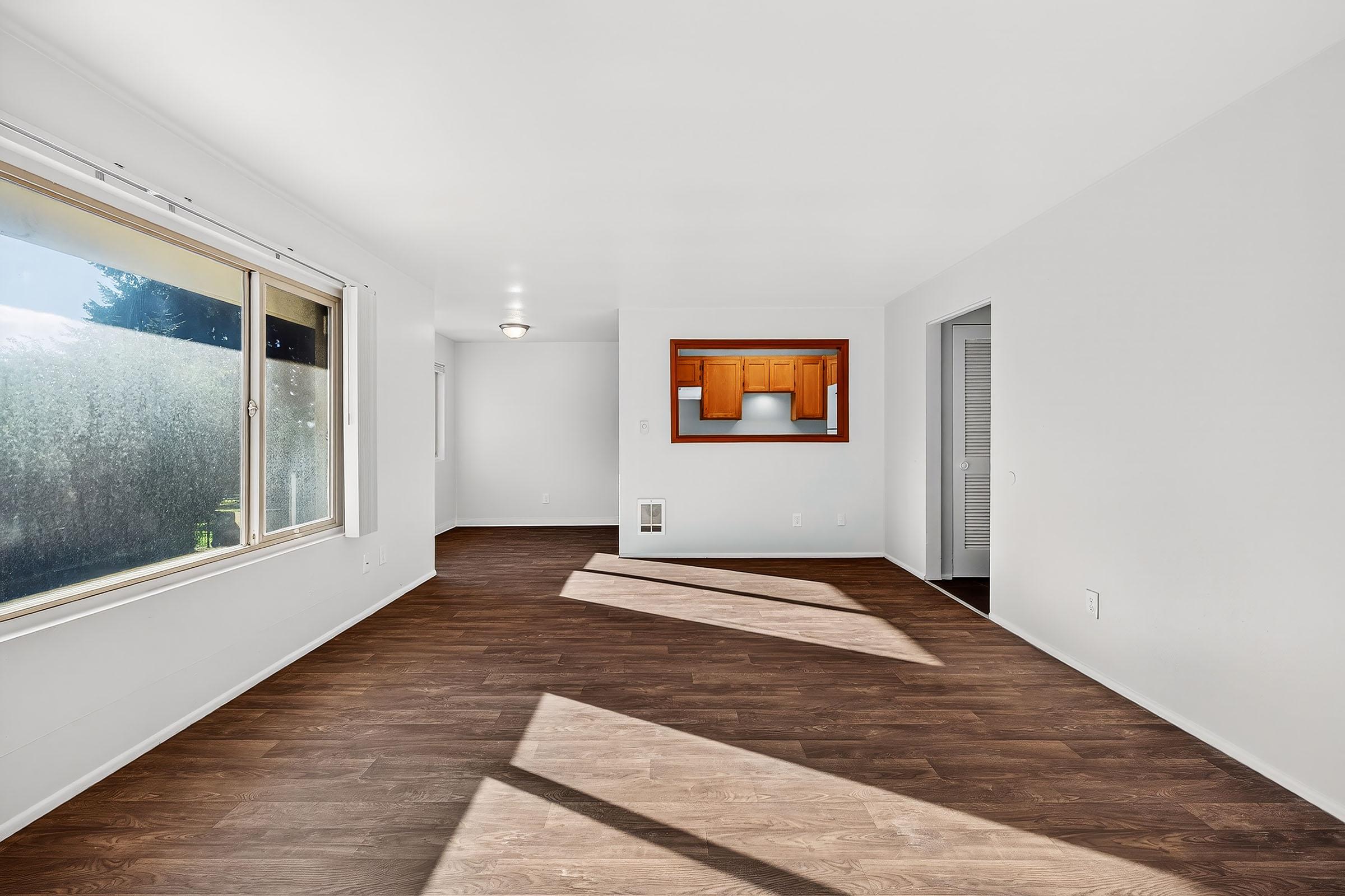 A bright, empty room with white walls and wooden flooring, featuring a large window on the left allowing natural light to enter. There is a small opening with a wooden bar counter in the center, leading to another room. Sunlight casts shadows on the floor, creating a warm atmosphere.