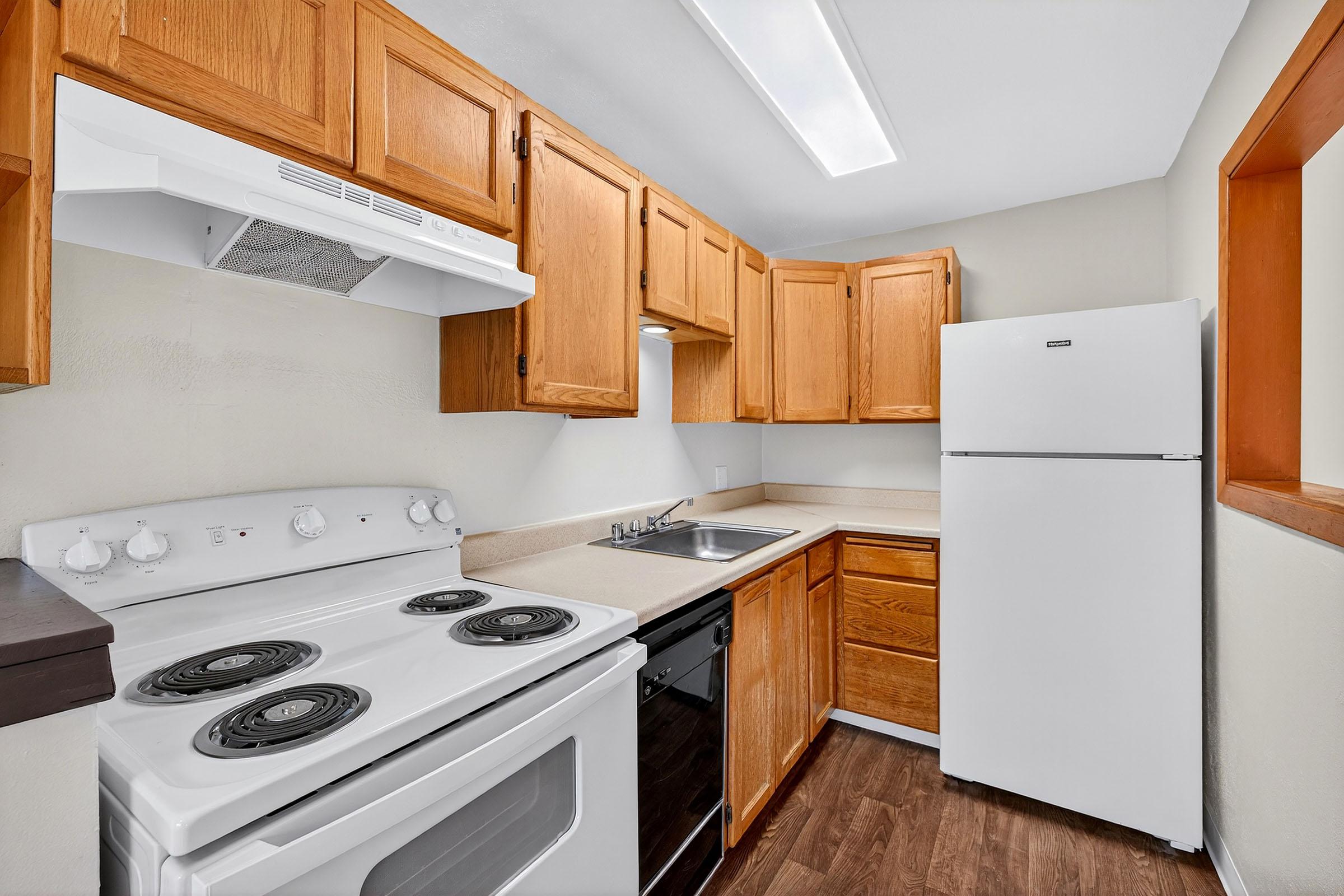 A modern kitchen featuring wooden cabinets, a white refrigerator, a stove with an oven, a sink, and a countertop. The space is well-lit with a ceiling light and has laminate flooring.