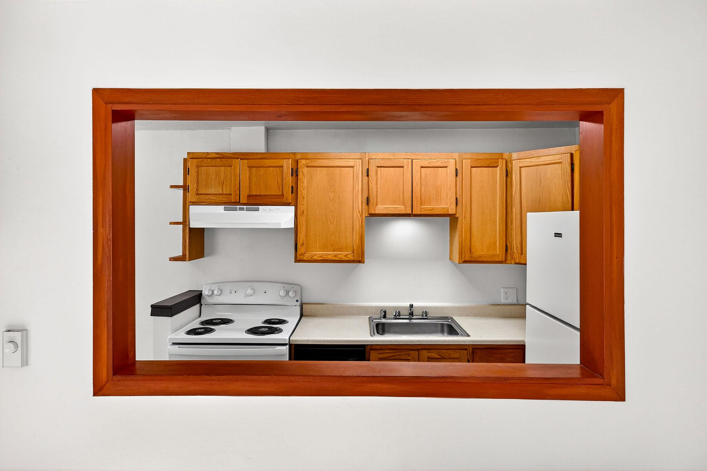 View of a kitchen through a framed opening. The kitchen features wooden cabinets, a white stove with four burners, a sink, and a refrigerator. The walls are painted white, and the space is well-lit, highlighting the counter and appliances.
