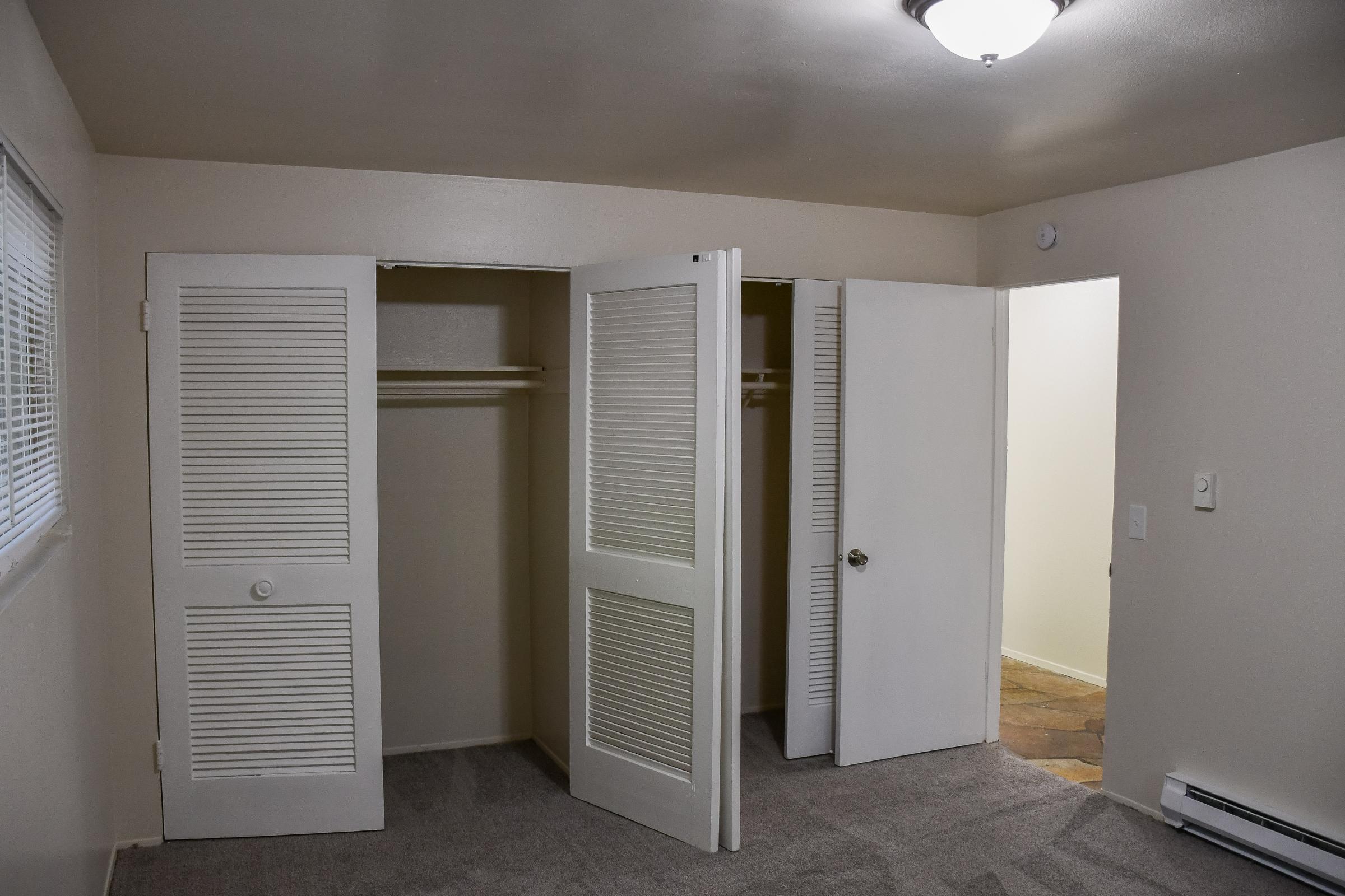 An empty room with light gray carpet and beige walls. There are two closed closet doors with louvered panels on one side, and a doorway leading to another area on the right. The ceiling light fixture is visible, illuminating the space. The room appears clean and well-maintained.