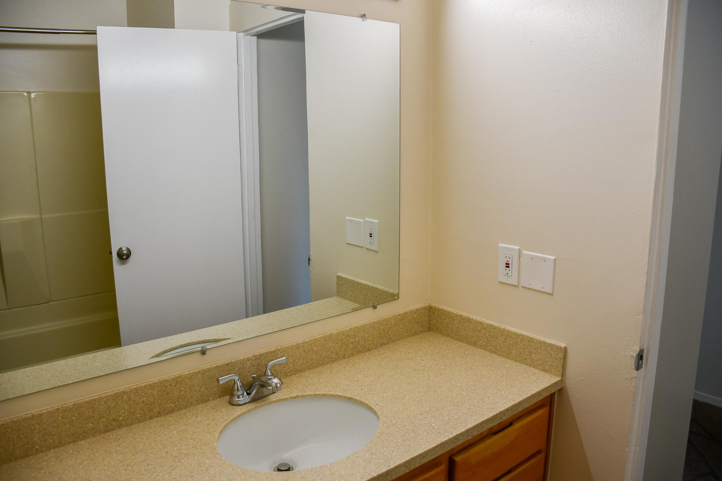 A bathroom scene featuring a countertop with a sink, a mirror above it, and a doorway leading to a shower area. The walls are painted a light color, and there are electrical switches on the wall next to the sink. The overall appearance is clean and simple.