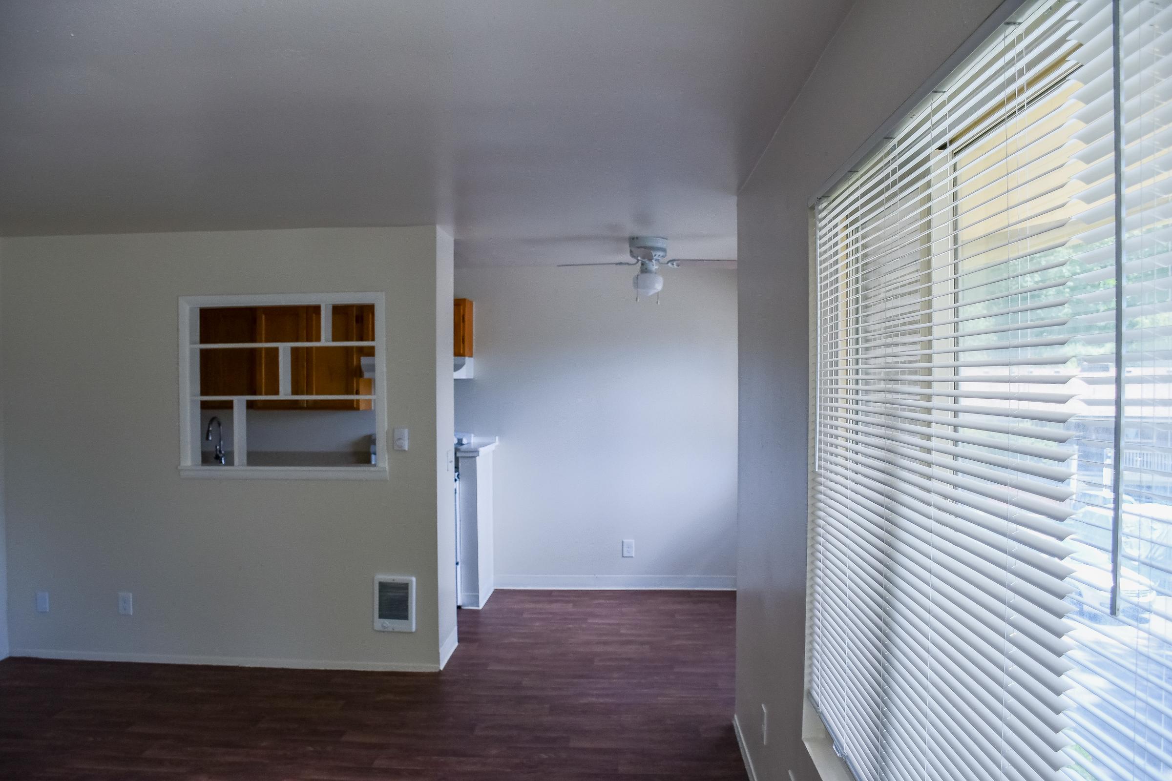A bright, spacious living area with light-colored walls and wooden flooring. To the left, there is a small kitchen area with wooden cabinets and appliances visible. On the right, a window with white blinds lets in natural light, and a ceiling fan is mounted above, enhancing the airy atmosphere.