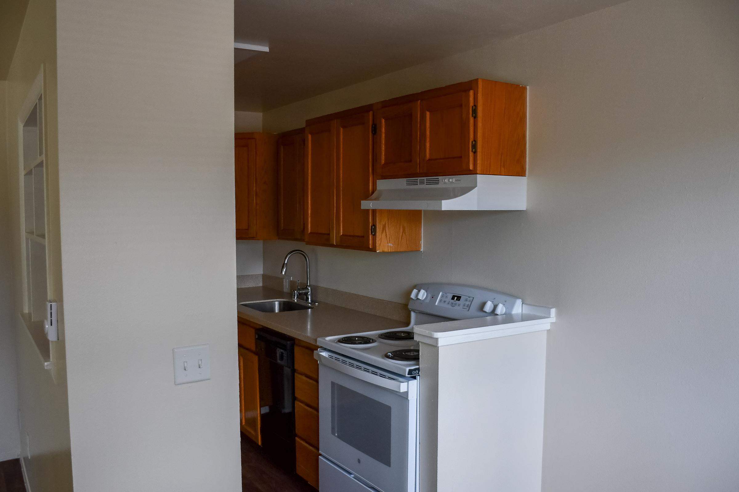 A small kitchen featuring wooden cabinets, a stainless steel sink, a stove, and an oven. The walls are painted in a light color, and there is a white range hood above the stove. The kitchen has a clean and modern appearance with a doorway leading to another room.