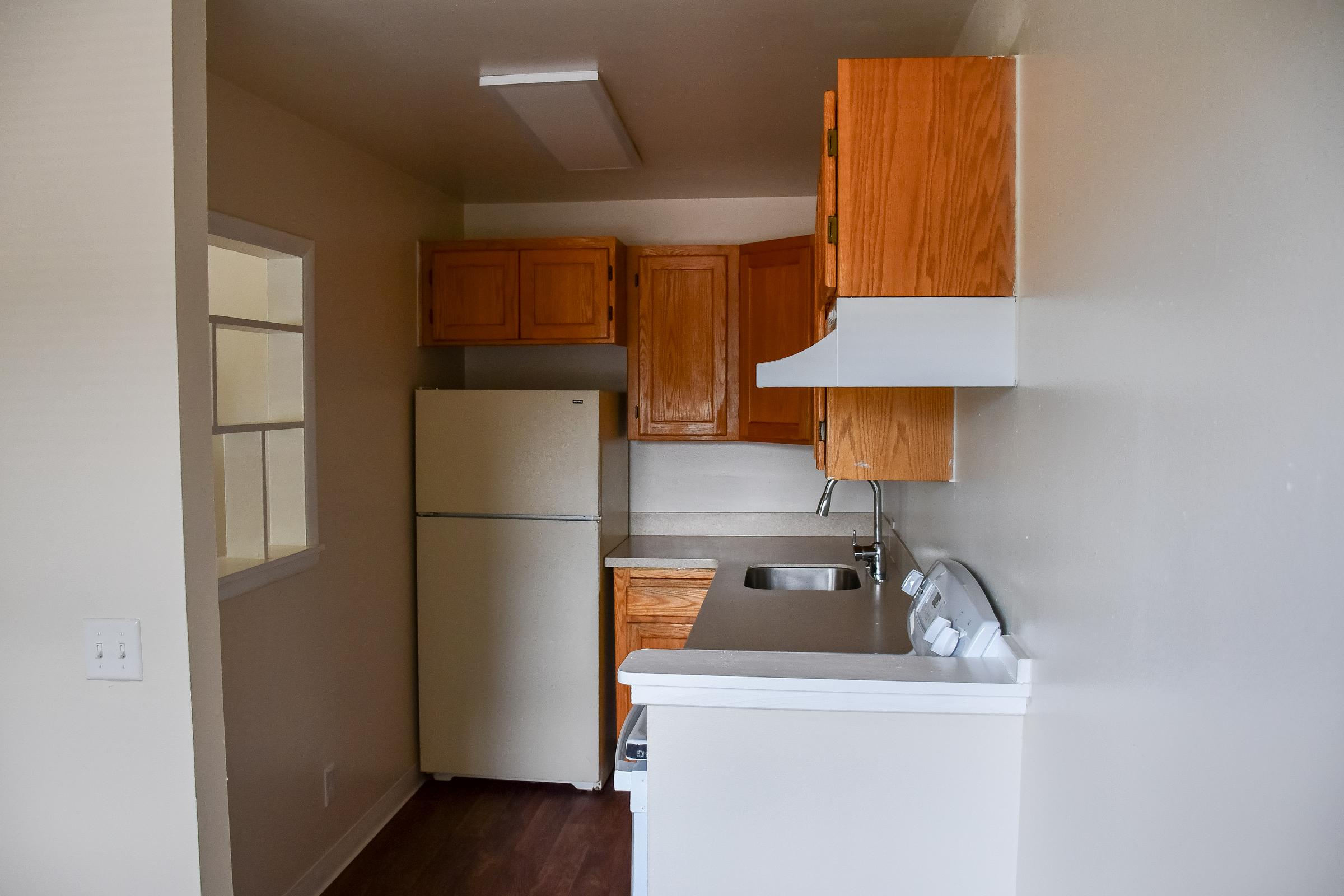 A compact kitchen featuring wooden cabinets, a white refrigerator, a sink, and a countertop with a stove and hood. Natural light enters through a nearby window, illuminating the space. The flooring is a dark, wooden design, contributing to a cozy atmosphere.