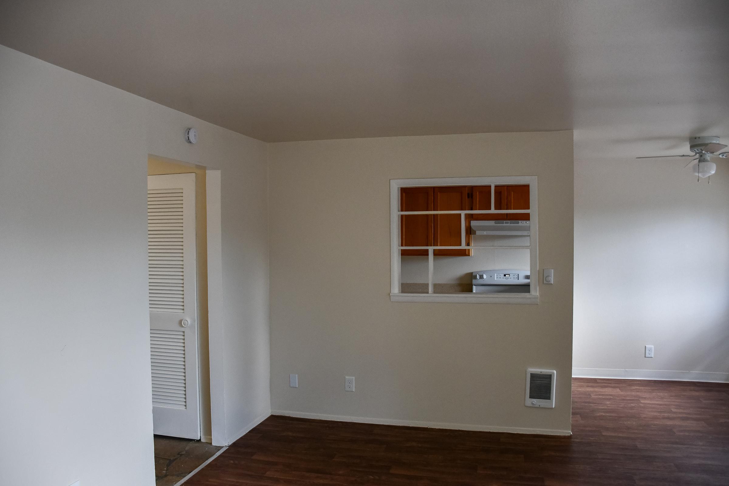Interior view of a small, light-colored living space showing an open layout. There is a kitchen area visible through a pass-through window, featuring wooden cabinets and a stove. A door on the left leads to another room. The flooring is dark wood, and the ceiling has a light fixture and ceiling fan.