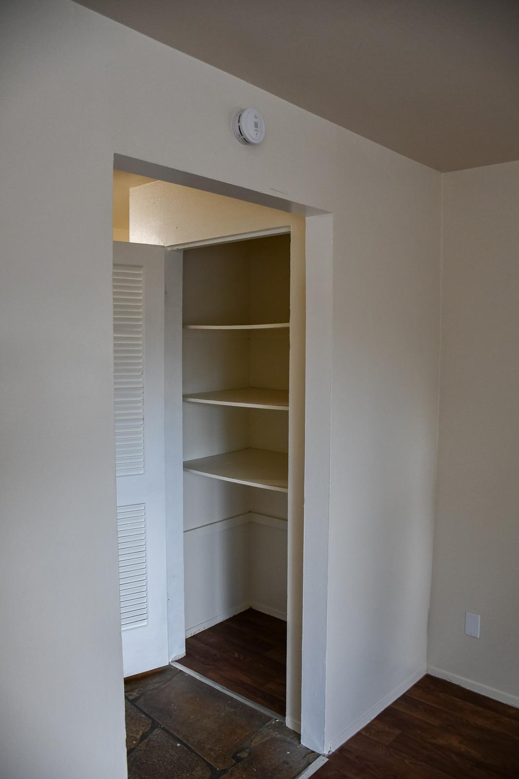 A small, empty closet with shelves along the back wall. The walls are painted a light color, and the closet entrance is framed by a plain white opening. The floor features a combination of stone and wood textures, with a smoke detector mounted on the ceiling.