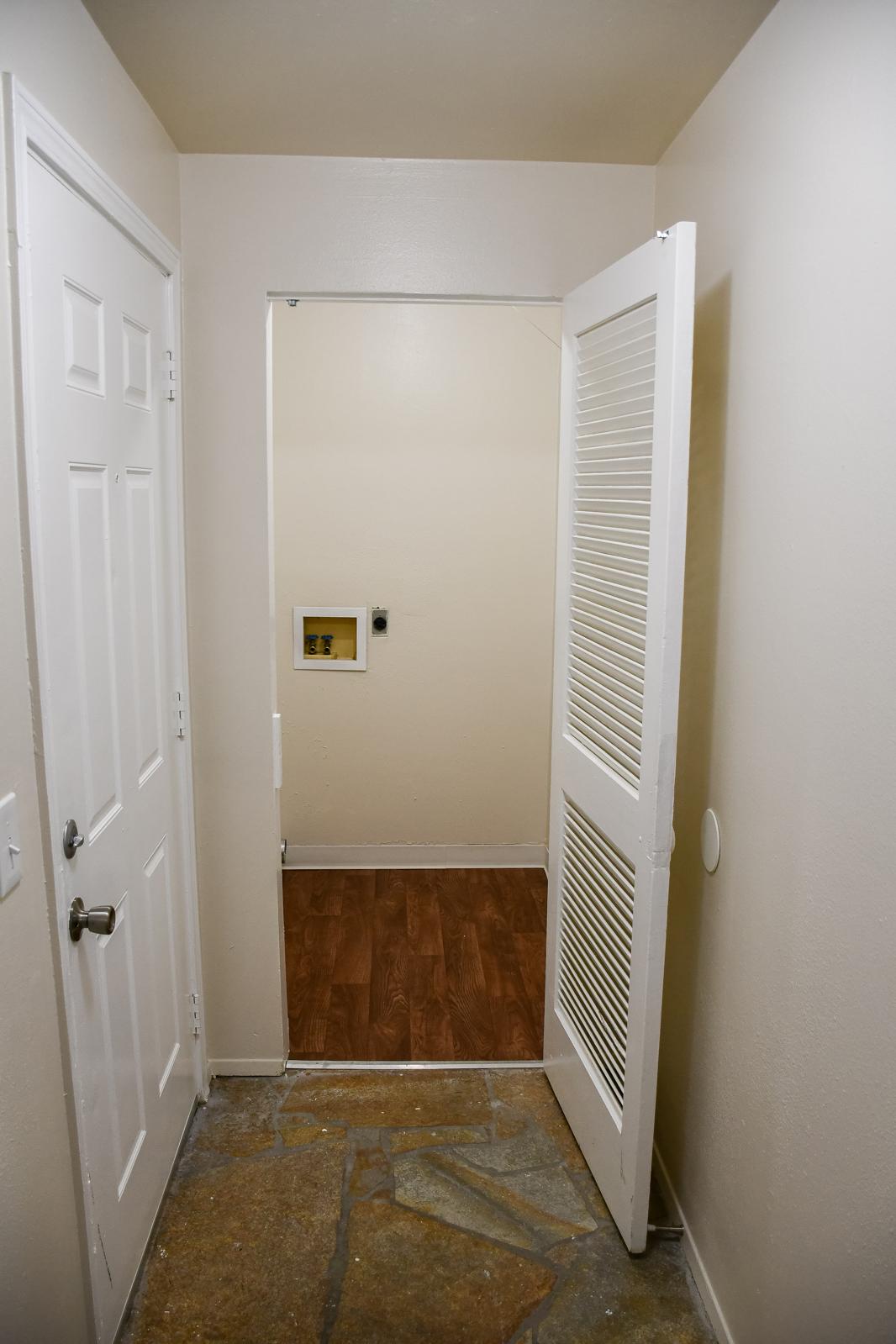A view of a hallway featuring a white door on the left and an open white louvered door leading into a small room. The walls are painted a light color, and the flooring is a mix of tile and wood. There's an electrical outlet visible on the wall in the small room.