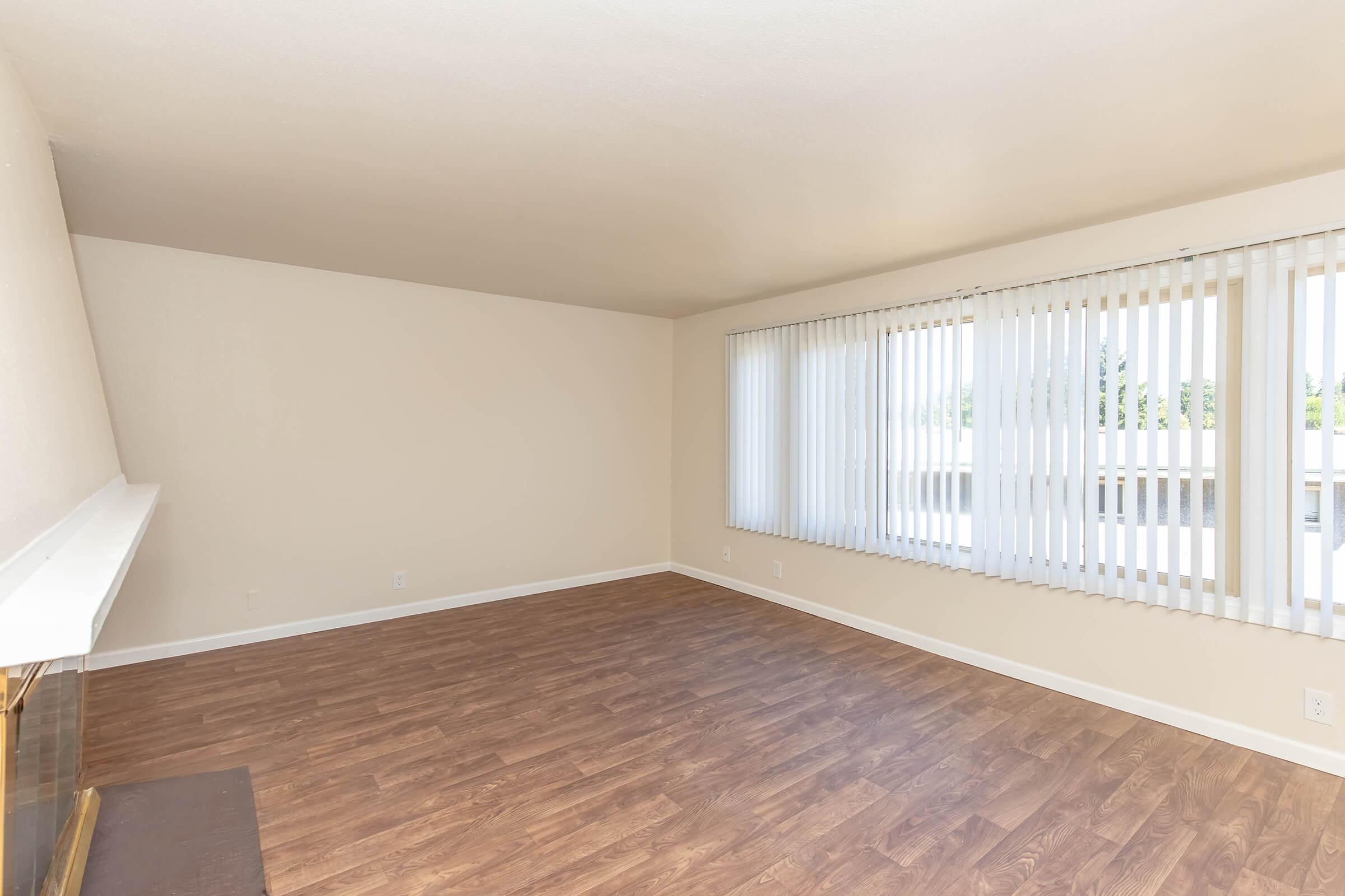Empty room featuring light-colored walls, a large window with vertical blinds allowing natural light to enter, and a wooden floor. A decorative fireplace is visible along one wall, creating a cozy atmosphere. The space is bright and uncluttered, suitable for various interior design possibilities.
