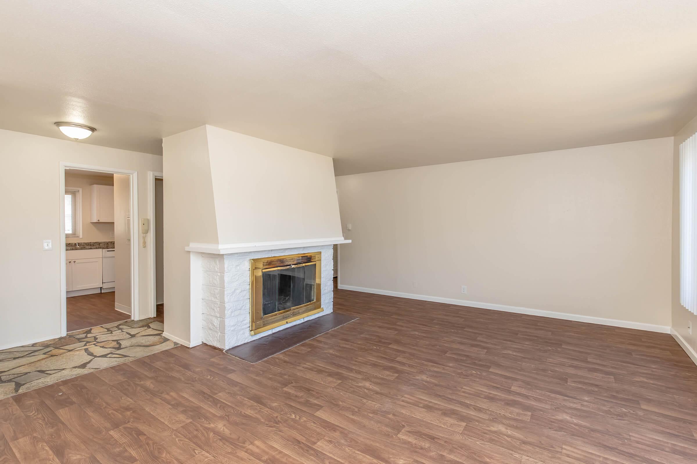 Spacious living room with a light-colored wall, hardwood floors, and a fireplace with a wooden mantel. A doorway on the left leads to a kitchen area, while large windows allow natural light to fill the room. The overall ambiance is bright and inviting.