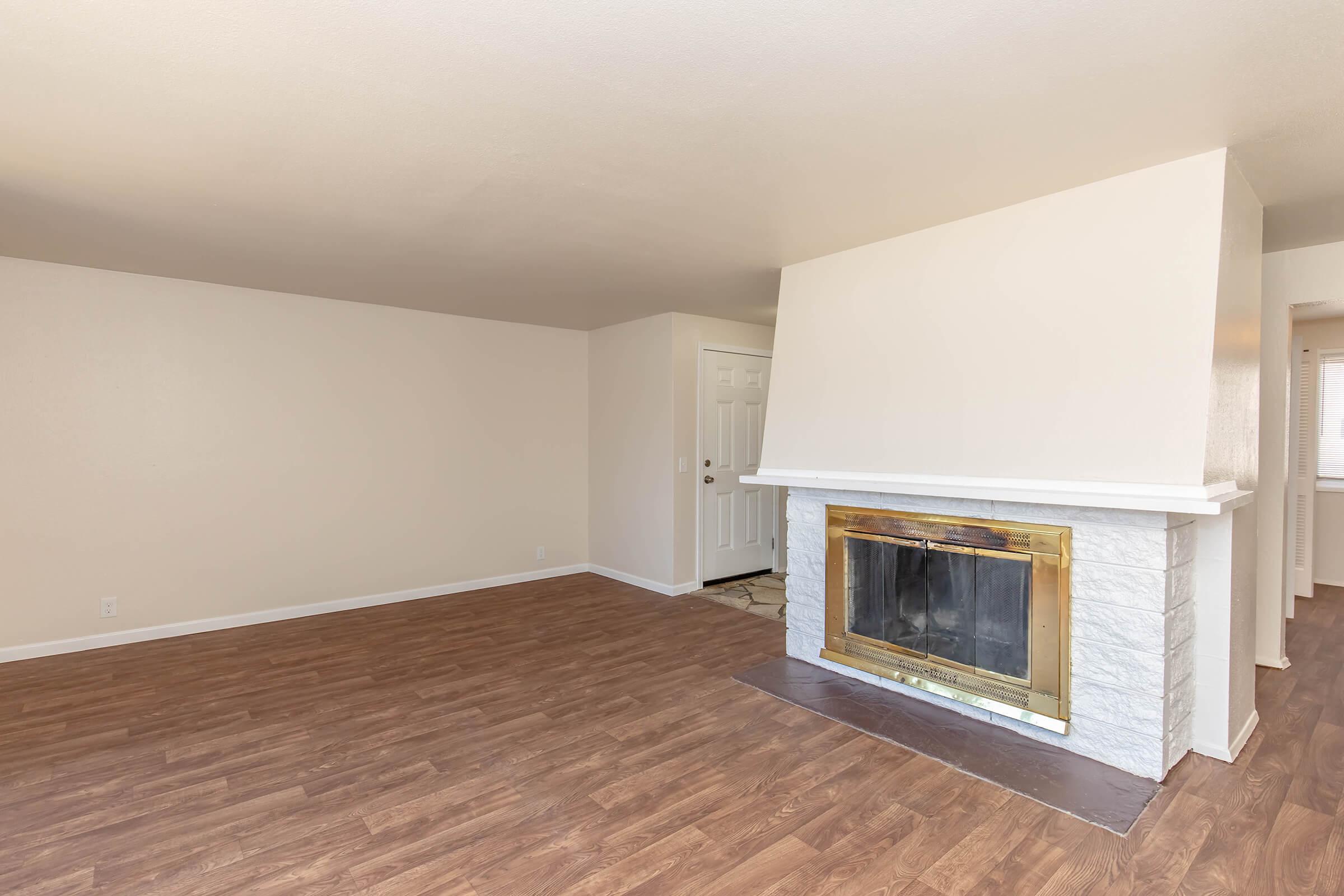 A spacious living room featuring a fireplace with a golden trim, light-colored walls, and a brown laminate floor. There's a white door on the left leading to another room, and the right side shows an entryway to another space. The room is well-lit with natural light.
