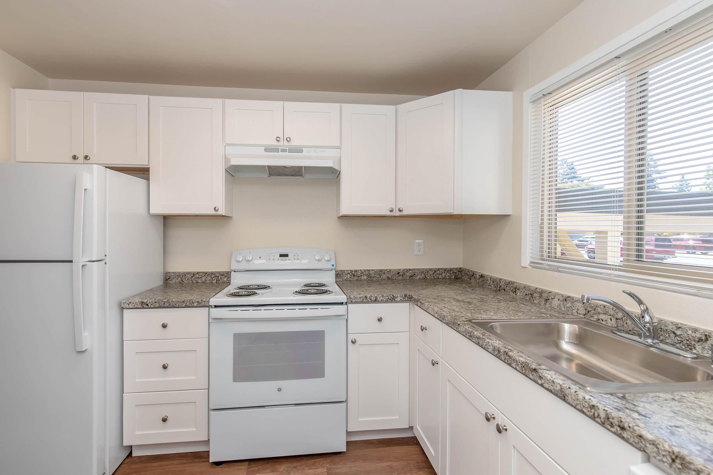 A modern kitchen featuring white cabinetry, a stainless steel sink, granite countertops, an electric stove with an overhead microwave, and a white refrigerator. Natural light comes through a window with blinds, illuminating the space. The floor appears to be wood or laminate.