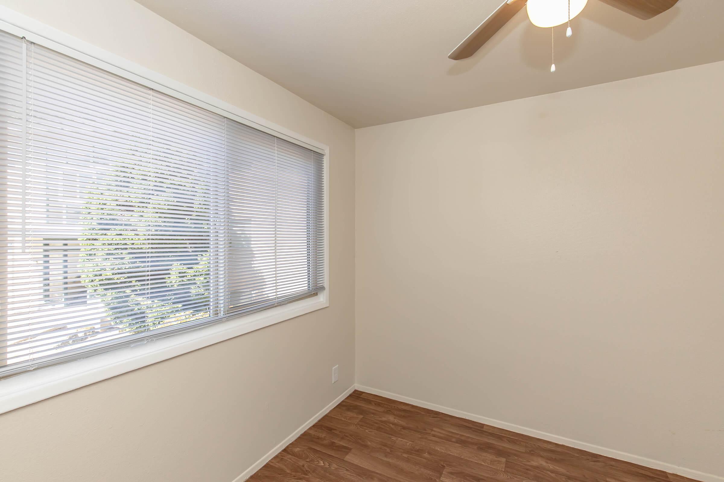 A small, empty room with light-colored walls and a window featuring white blinds. The floor is made of wooden laminate, and a ceiling fan is visible in the corner. Natural light is coming through the window, illuminating the space.