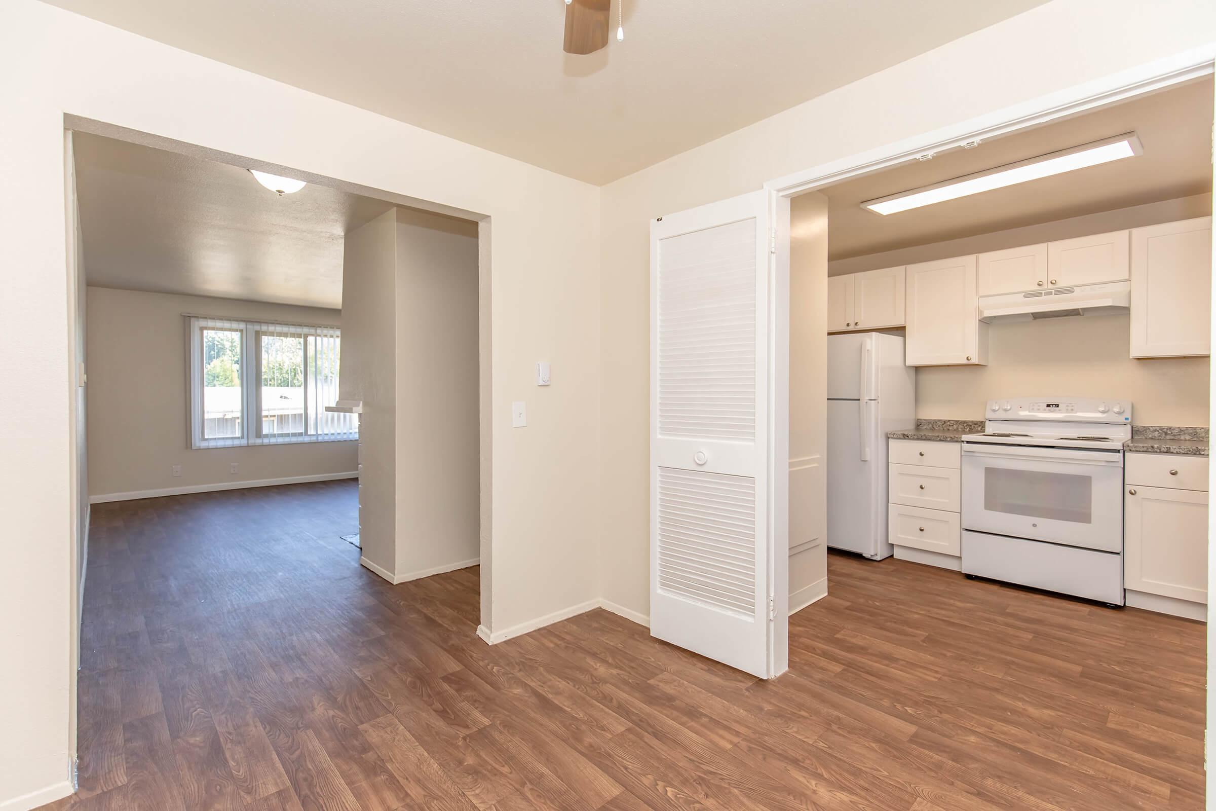 Bright, vacant apartment interior featuring a spacious living area with windows, and an adjoining kitchen equipped with white cabinetry, a refrigerator, and a stove. The floors are finished with wood-like laminate, and the walls are painted in neutral tones, creating a welcoming atmosphere.