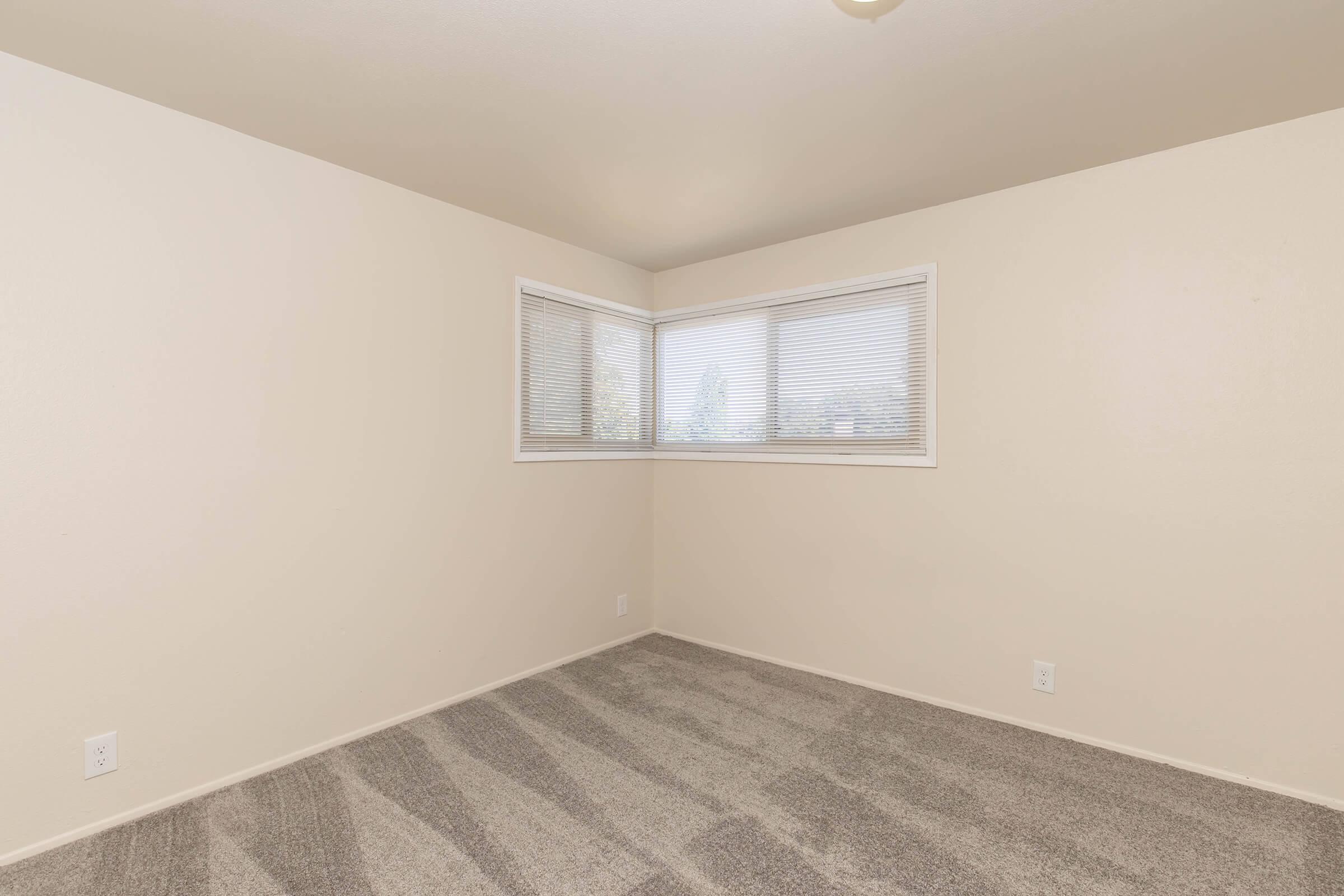 Empty room with light beige walls and carpet. A large window on one wall features white blinds, allowing natural light to enter. The space has no furniture, creating a minimalistic atmosphere.