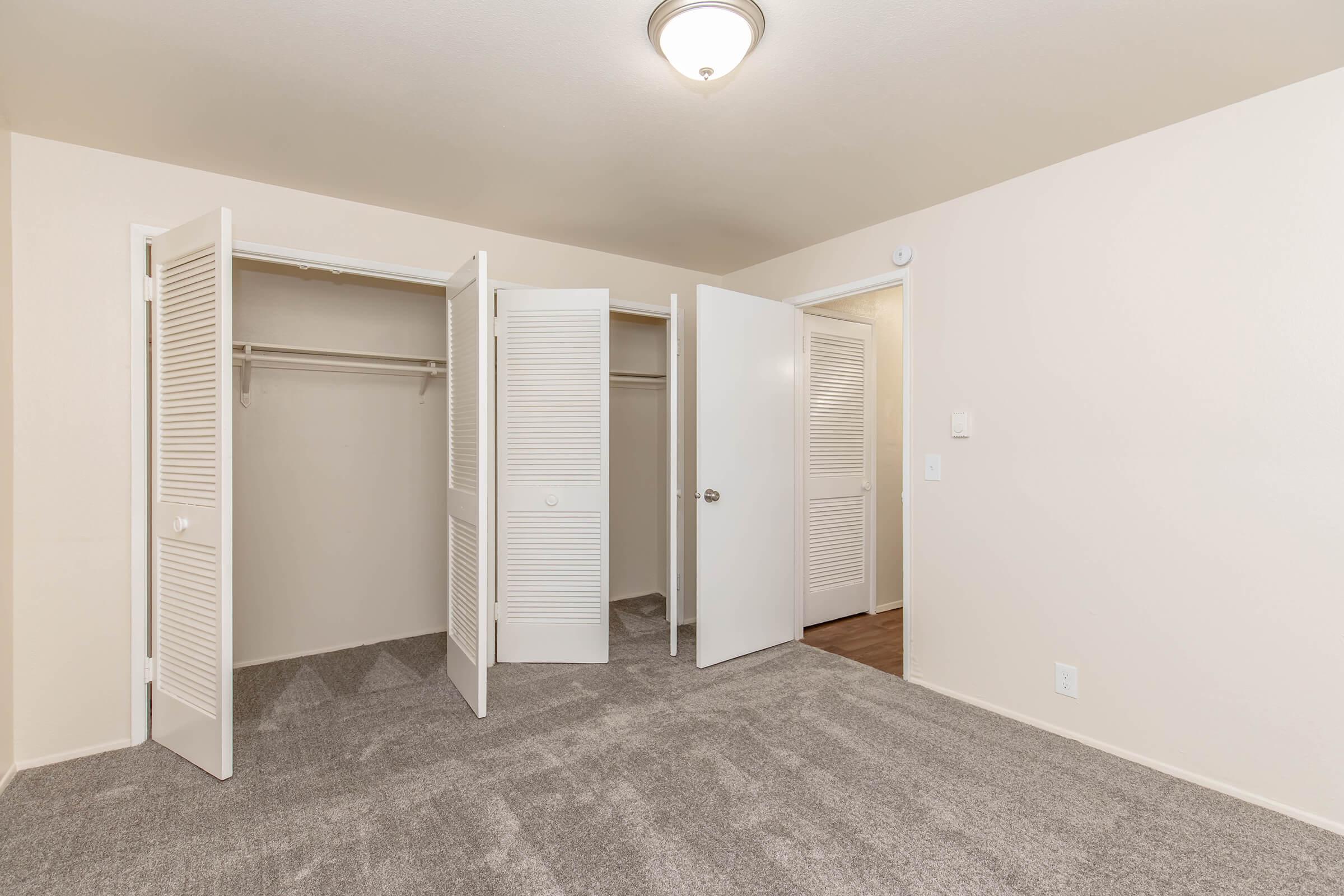 A well-lit bedroom featuring beige walls, plush gray carpet, and multiple white-paneled closet doors. The room is empty, showcasing ample space with a ceiling light fixture and a neutral aesthetic, ideal for decorating or moving in.
