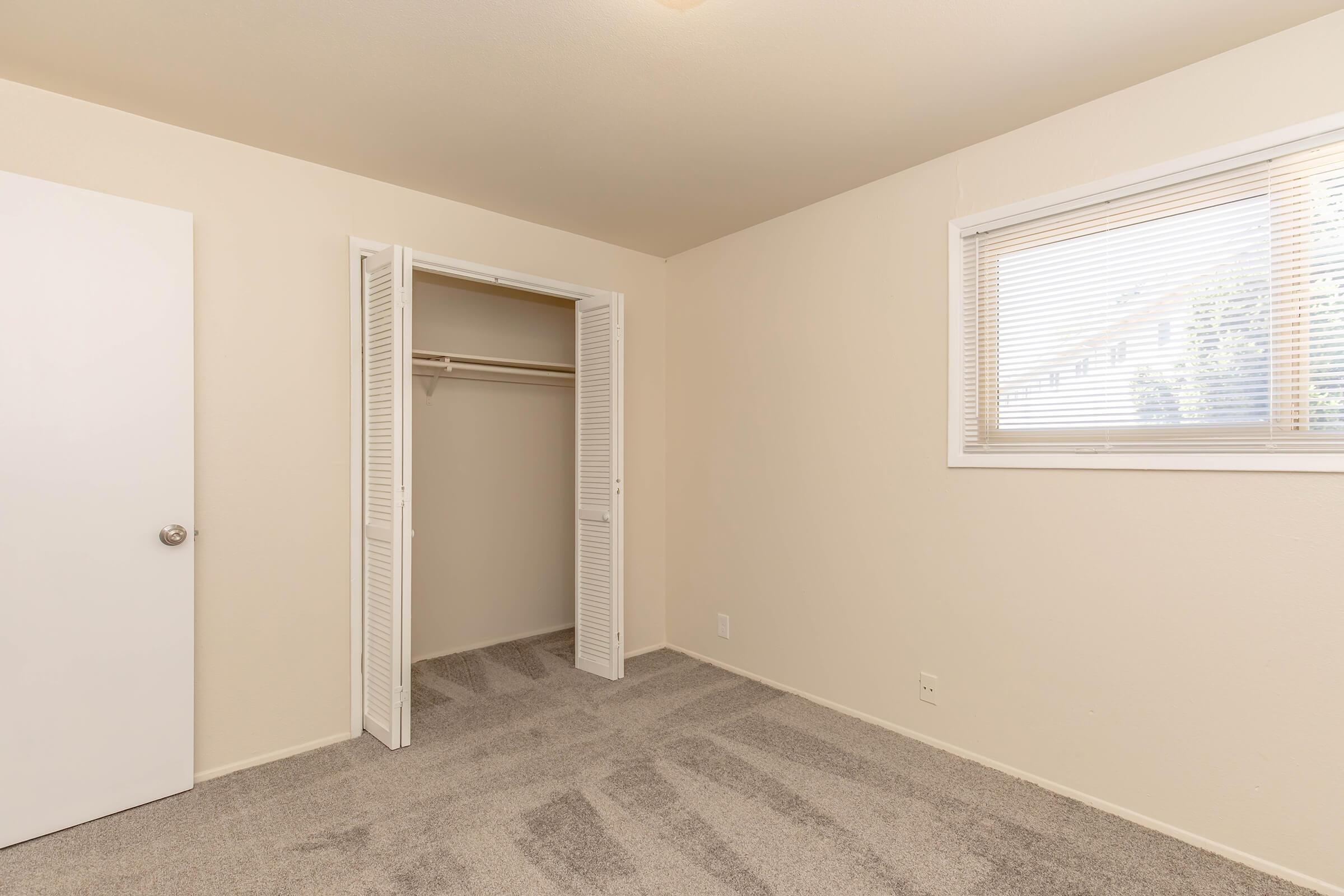 A vacant room with light beige walls and gray carpet. There's a white door leading to a closet with dual sliding doors. A window with blinds allows natural light in, and the overall space appears well-lit and tidy.