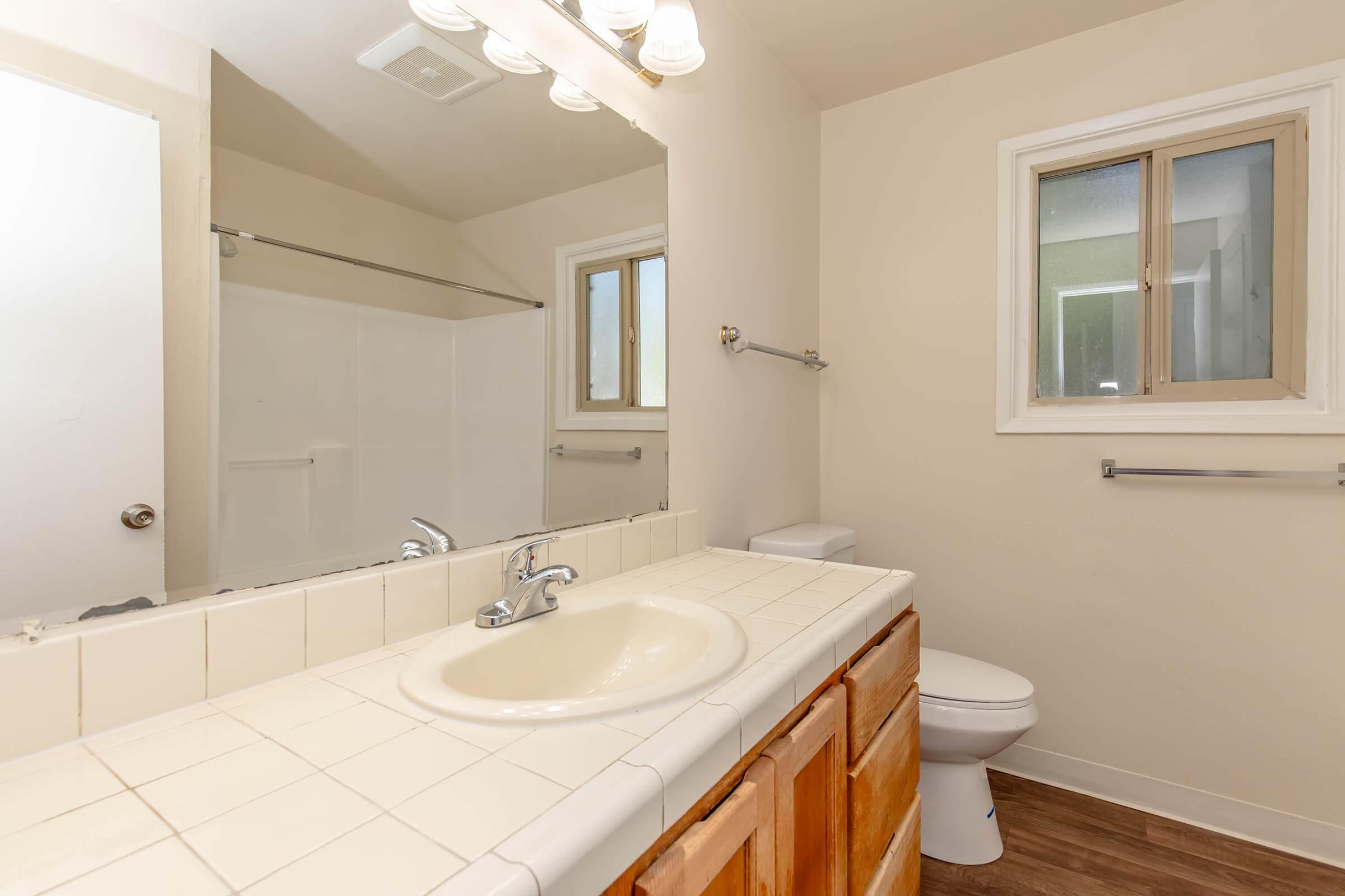A bright and clean bathroom featuring a sink with a mirror above, wooden cabinetry, a toilet, and a shower area. Natural light comes through a window, and the walls are painted in a soft shade. The floor has a wood-like finish, creating a warm and inviting atmosphere.