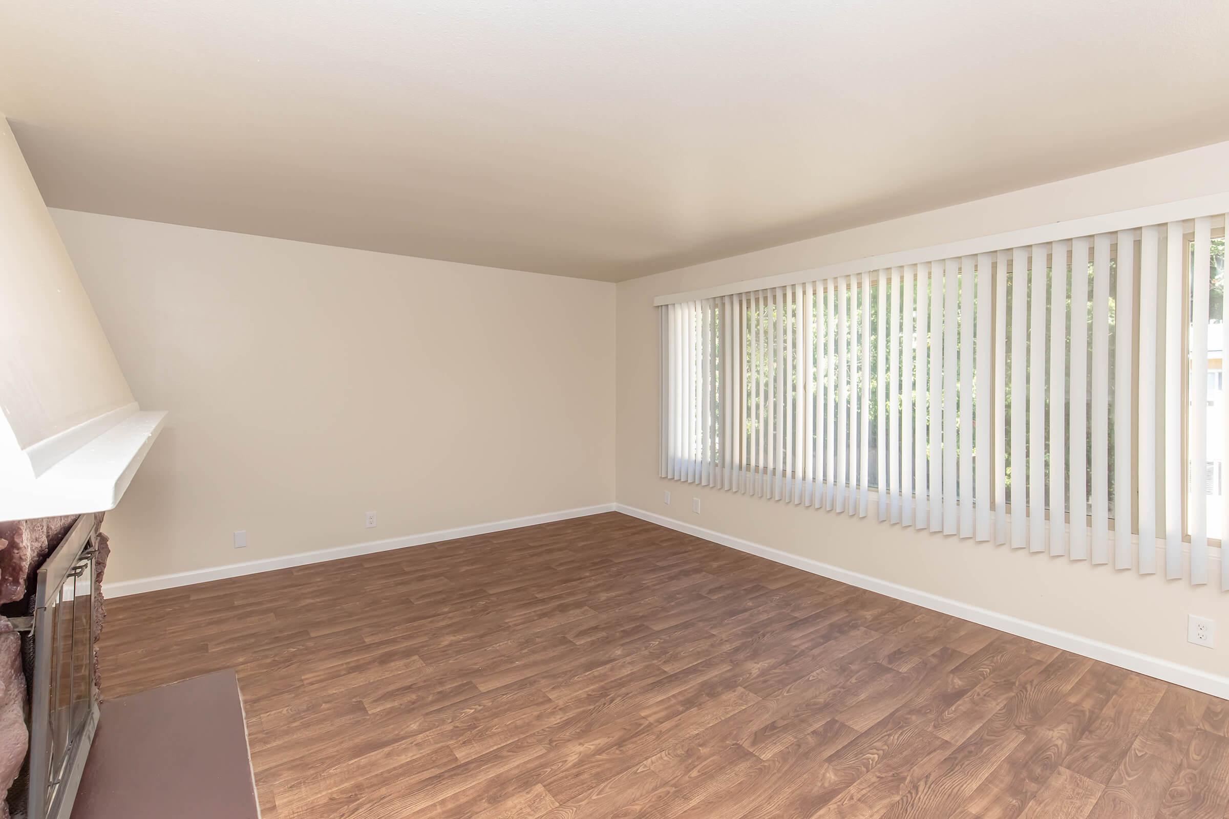 Empty room with light-colored walls and wooden flooring. Features a large window with vertical blinds allowing natural light, and a white-painted fireplace on one side. The space is uncluttered, creating a bright and open atmosphere, suitable for various interior design possibilities.