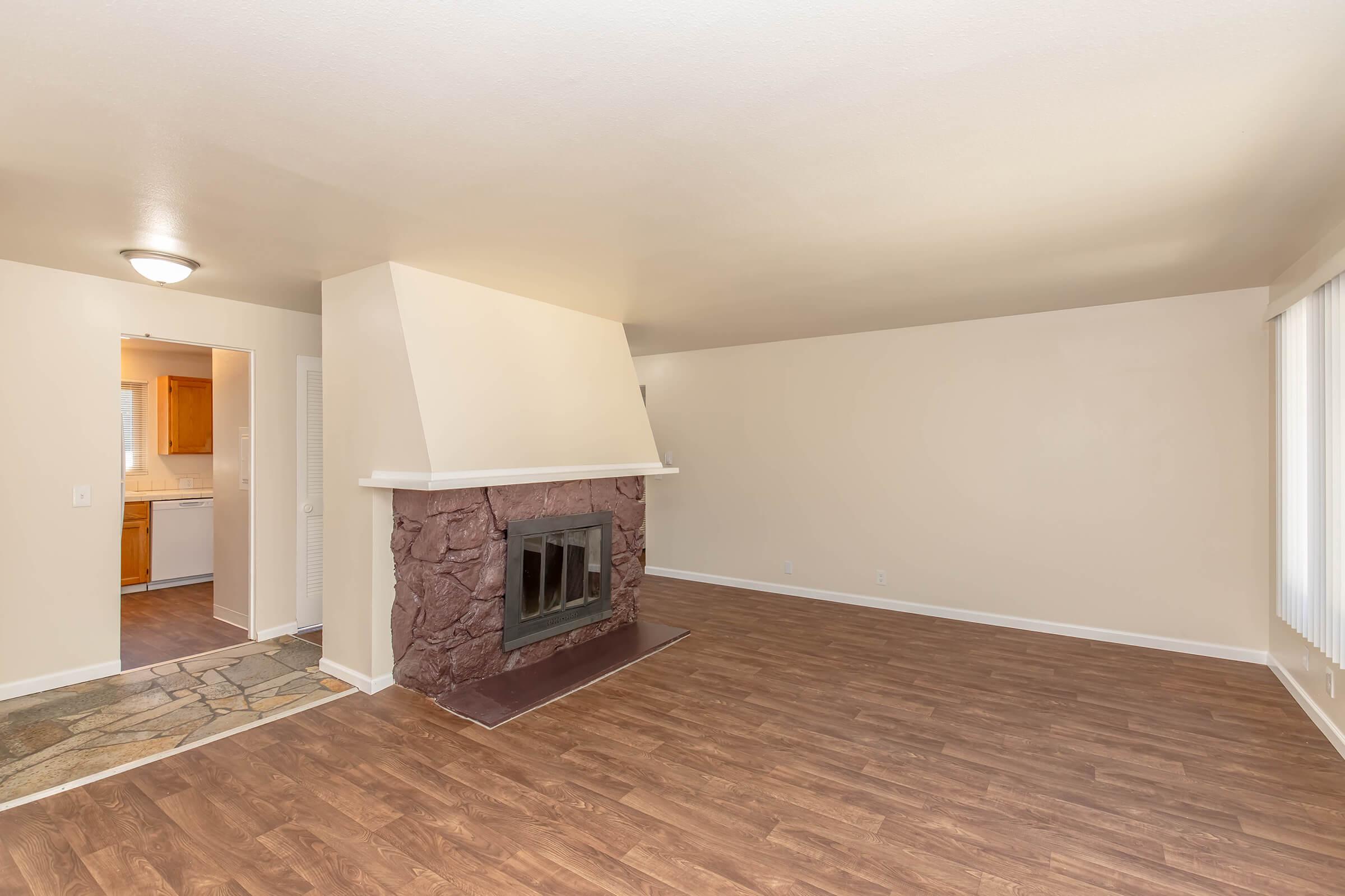 Spacious living room with light-colored walls and a stone fireplace as the focal point. Brown laminate flooring extends throughout the room. A doorway leads to a kitchen, and large windows allow natural light to fill the space.