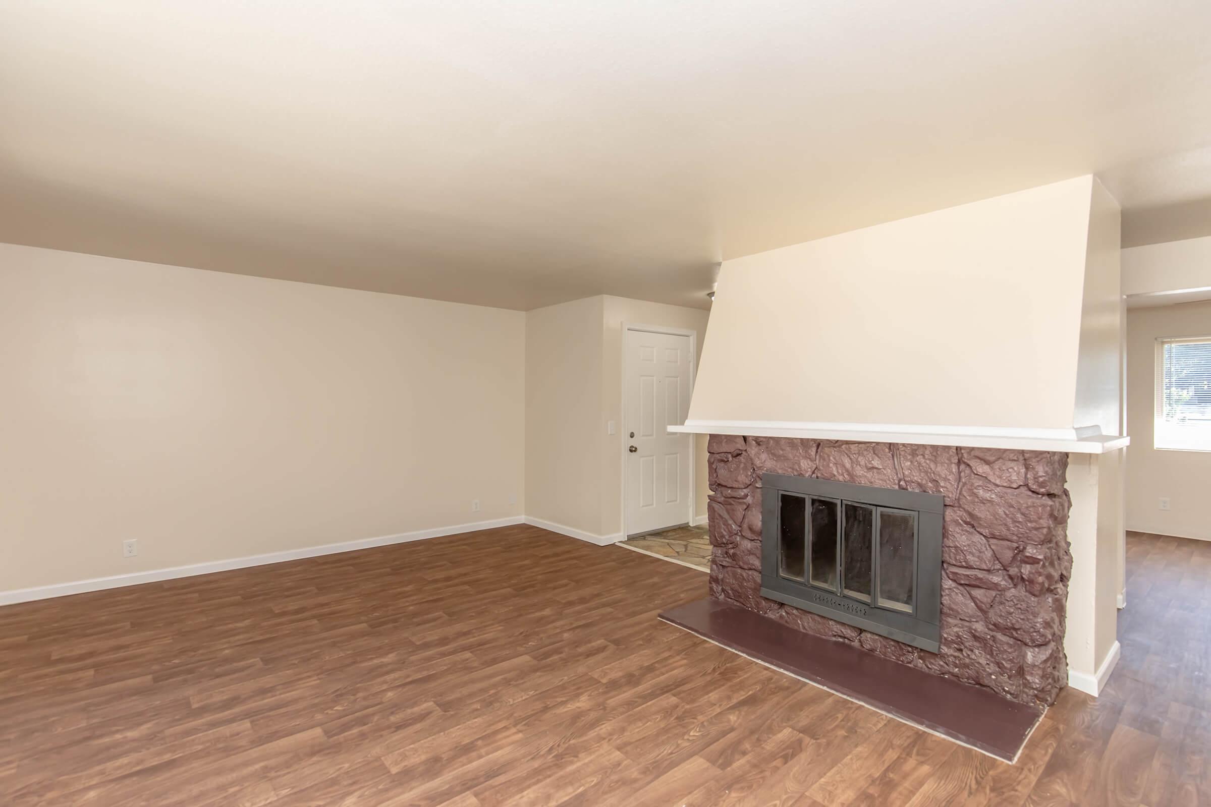 A spacious, empty living room featuring light beige walls and a large, stone fireplace with a dark mantle. The floor is finished with hardwood-like laminate, and there is a doorway visible in the background leading to another room. Natural light enters the space, creating a welcoming atmosphere.