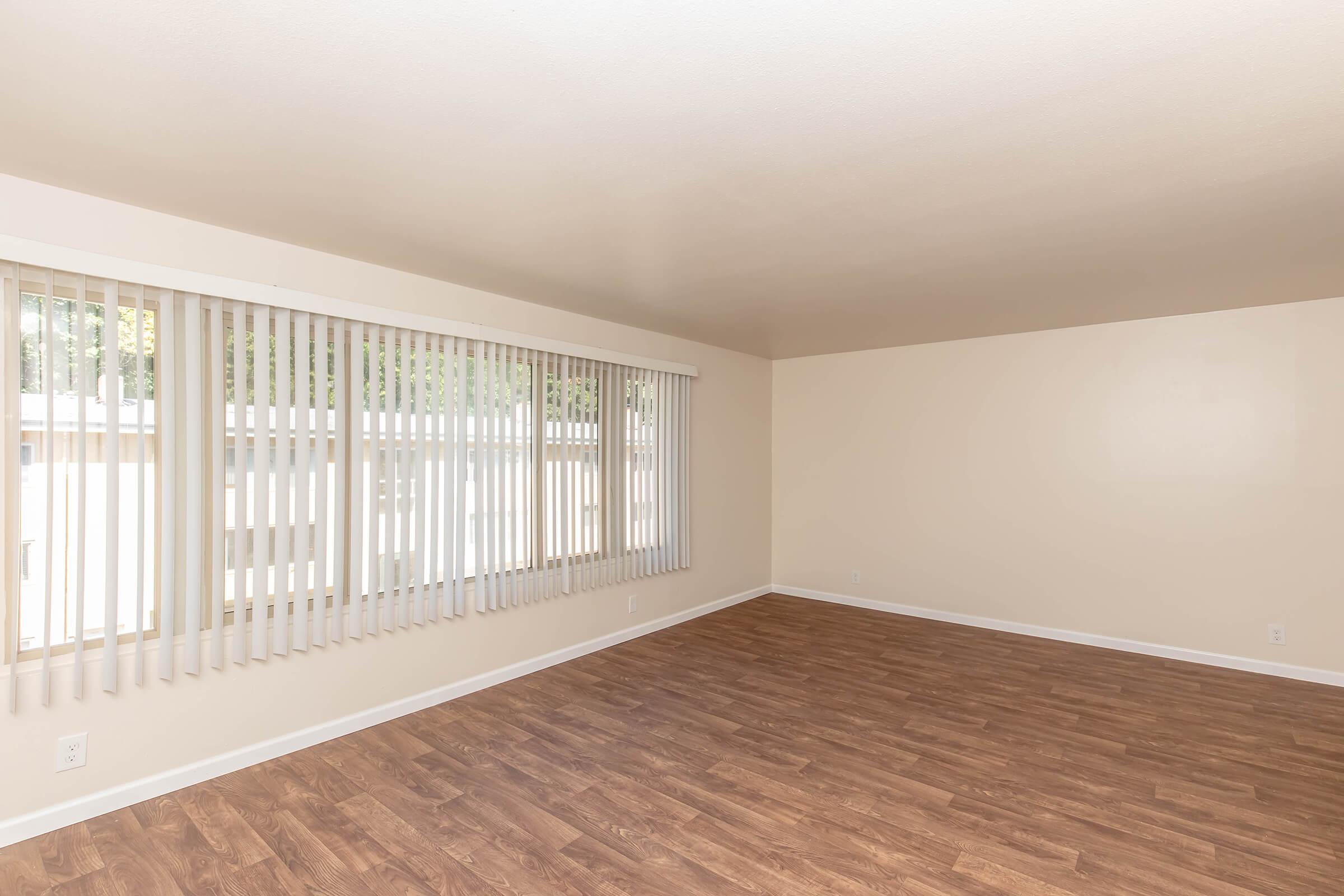 A spacious, empty room with light wood flooring and large windows covered by vertical blinds. The walls are painted a soft beige, giving the space a bright and airy feel. Natural light filters in, creating a calm and inviting atmosphere.