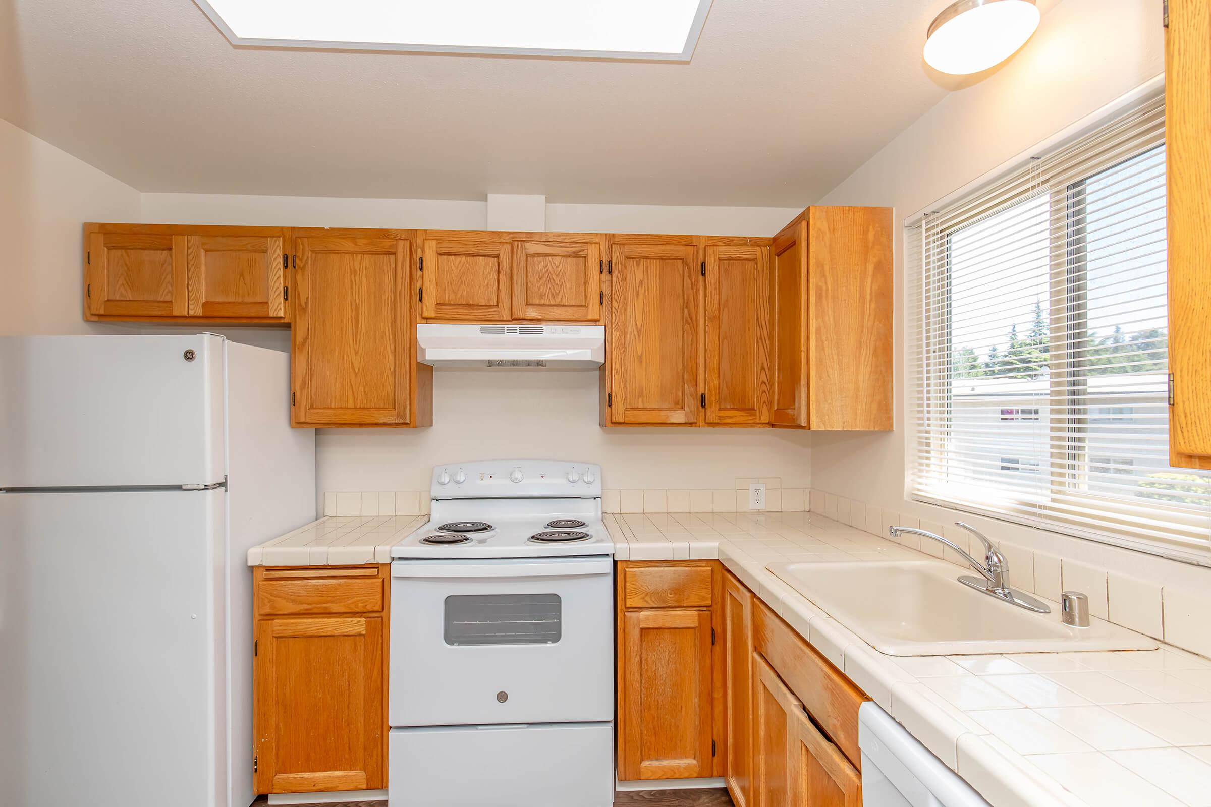 A kitchen featuring light wood cabinets, a white refrigerator, a white stove with an oven, and a sink. The counter is tile, and a window provides natural light. There is a range hood above the stove and blinds on the window.
