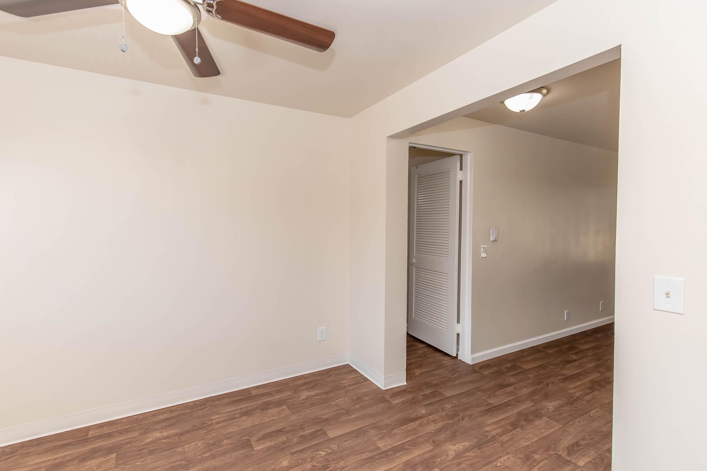 A well-lit, empty room featuring light brown wooden flooring, white walls, and a ceiling fan. An open doorway leads to another area, suggesting a spacious layout. The room is clean and offers a neutral, minimalist aesthetic.