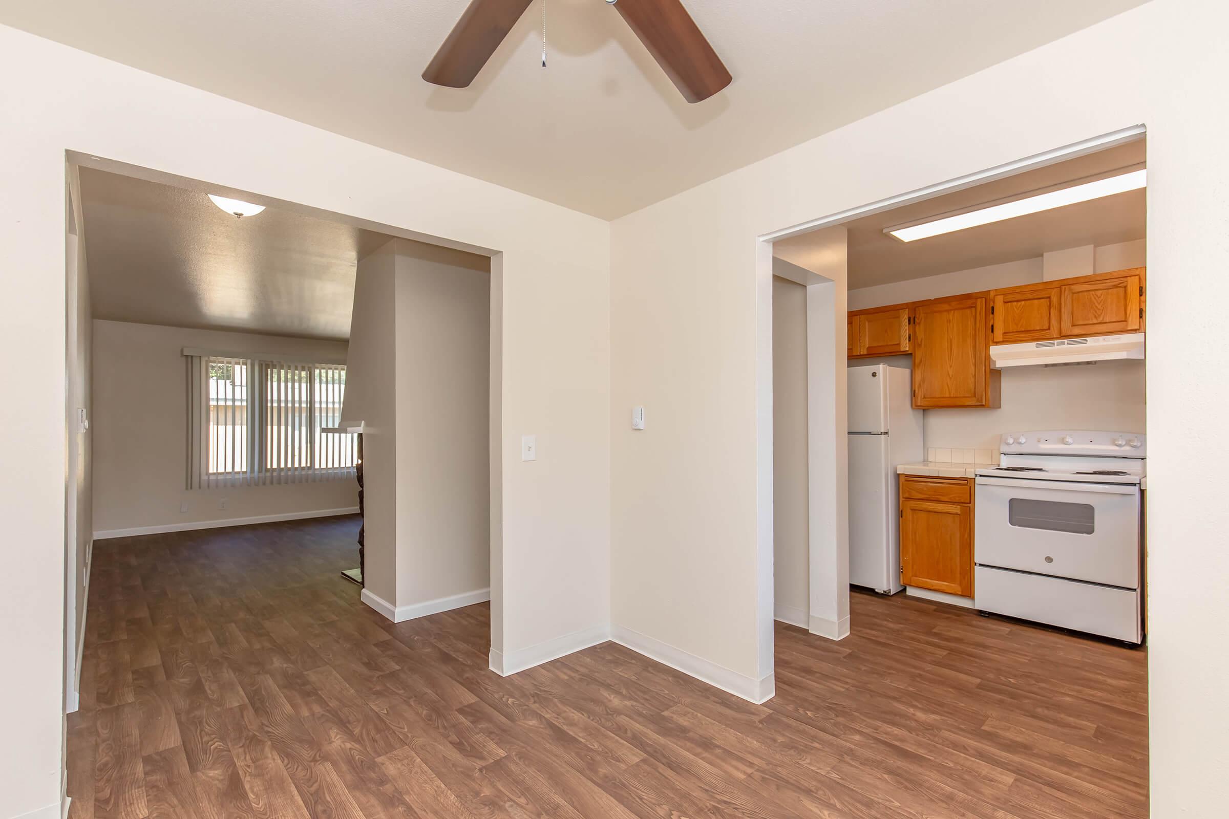 Interior view of a home featuring a living area to the left with large windows and wooden flooring, and a kitchen to the right with wooden cabinets, a white stove, and a refrigerator. A ceiling fan is visible above, and the overall design is bright and open.