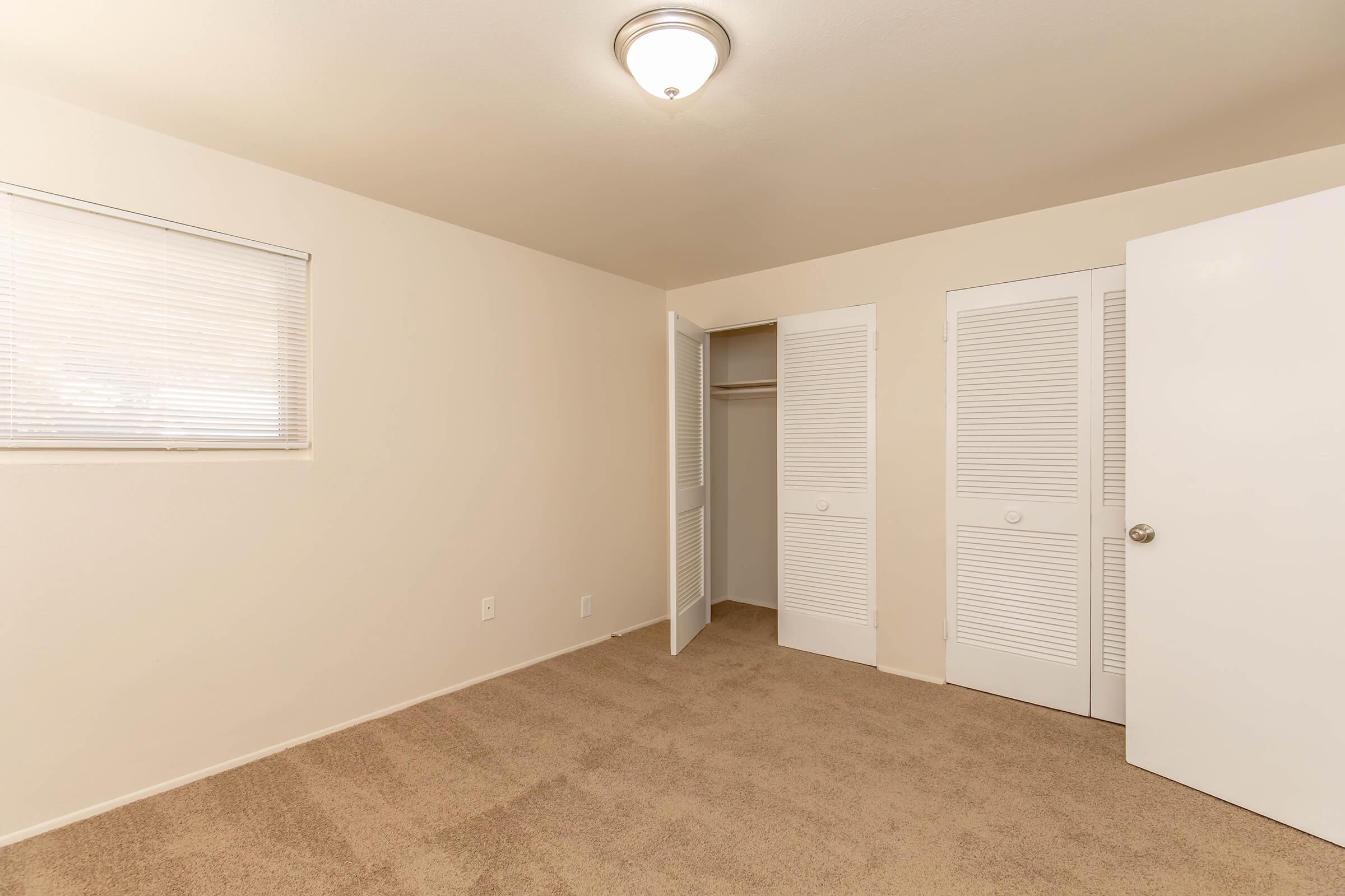 A vacant room with beige walls and carpet. There is a window with blinds on the left side and a closet with double doors on the right. The ceiling light fixture is visible, and the room appears spacious and well-lit.
