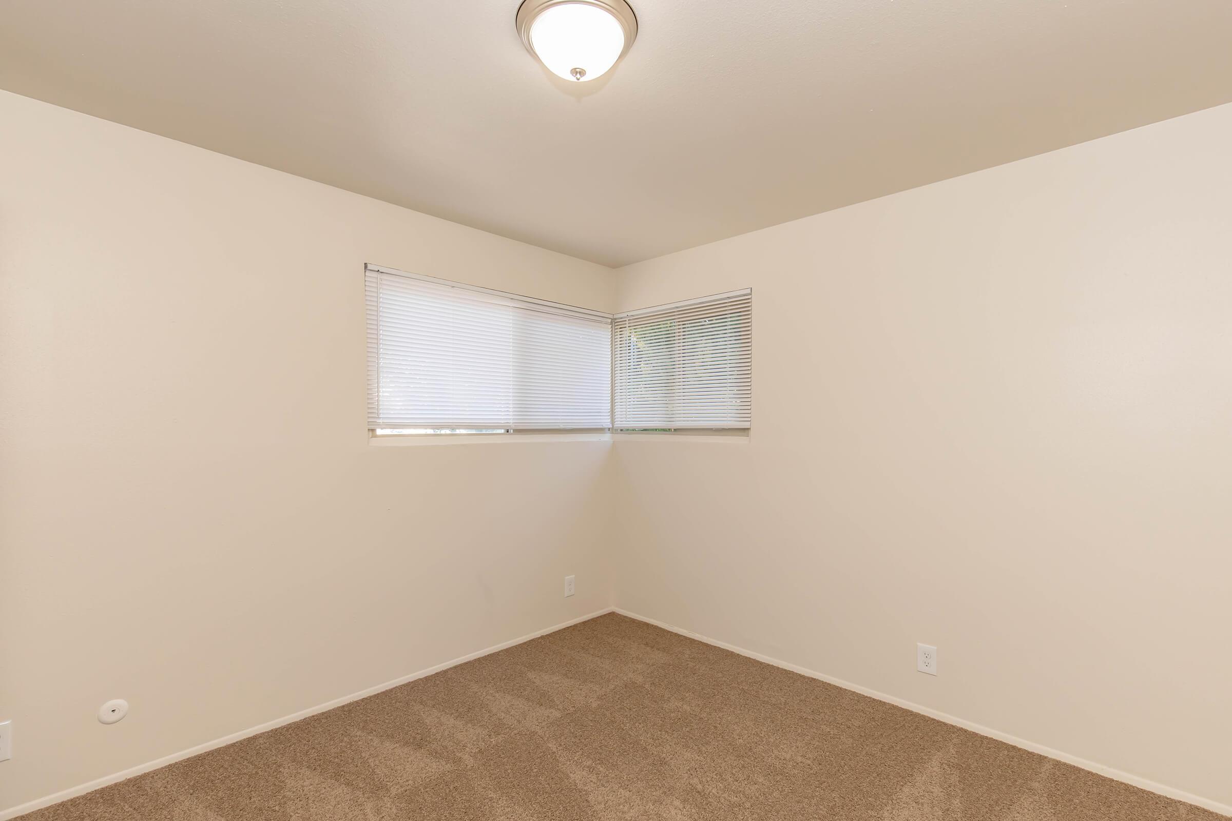 Empty room with beige walls and light brown carpet. There are two small windows with blinds providing natural light. The ceiling features a simple light fixture. The space is uncluttered and ready for furnishing.