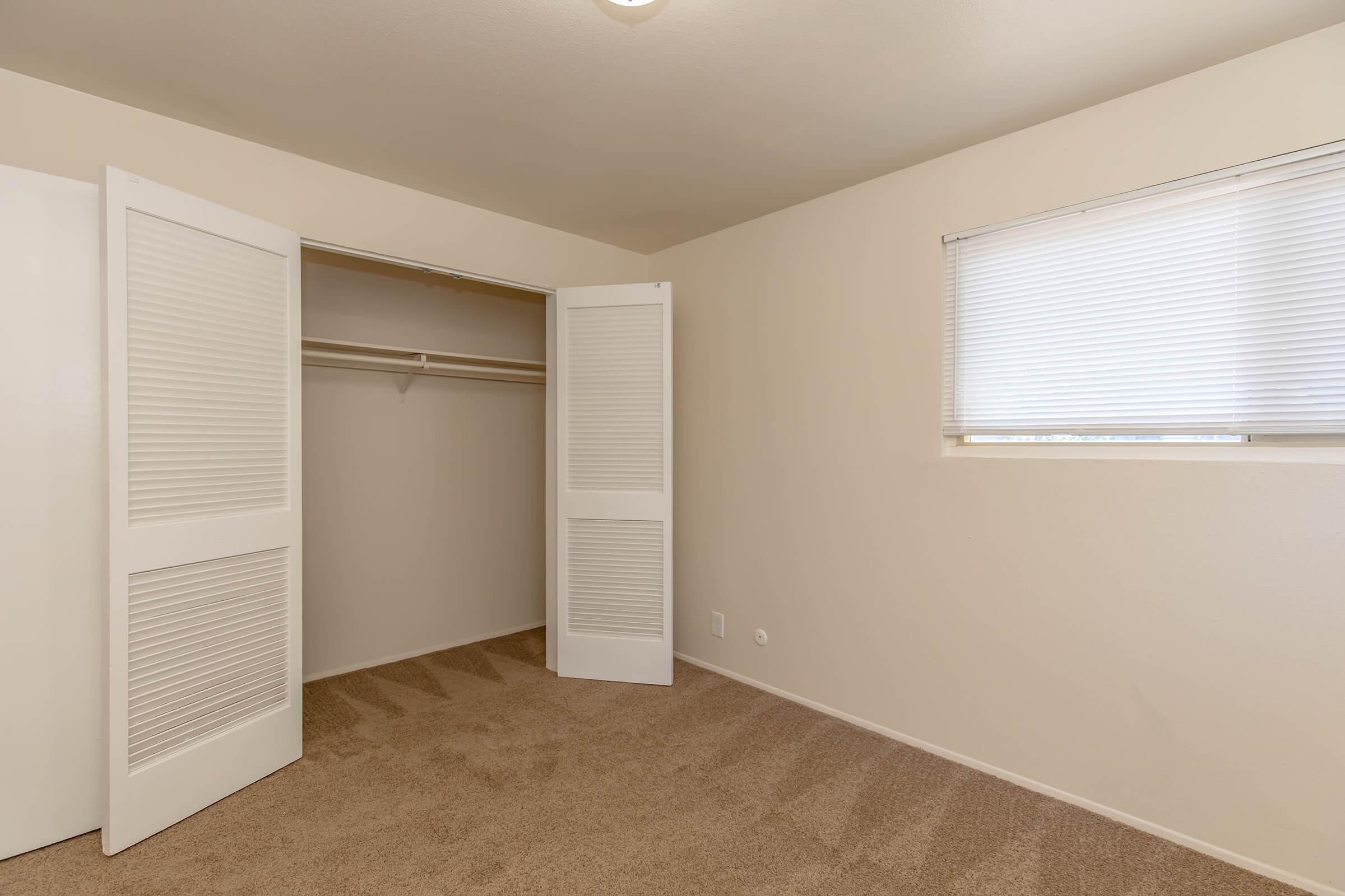 Empty room featuring beige walls and carpet, with a window dressed in blinds. A closet with white louvered doors is open, revealing an empty interior. The room is well-lit, providing a spacious and clean appearance.