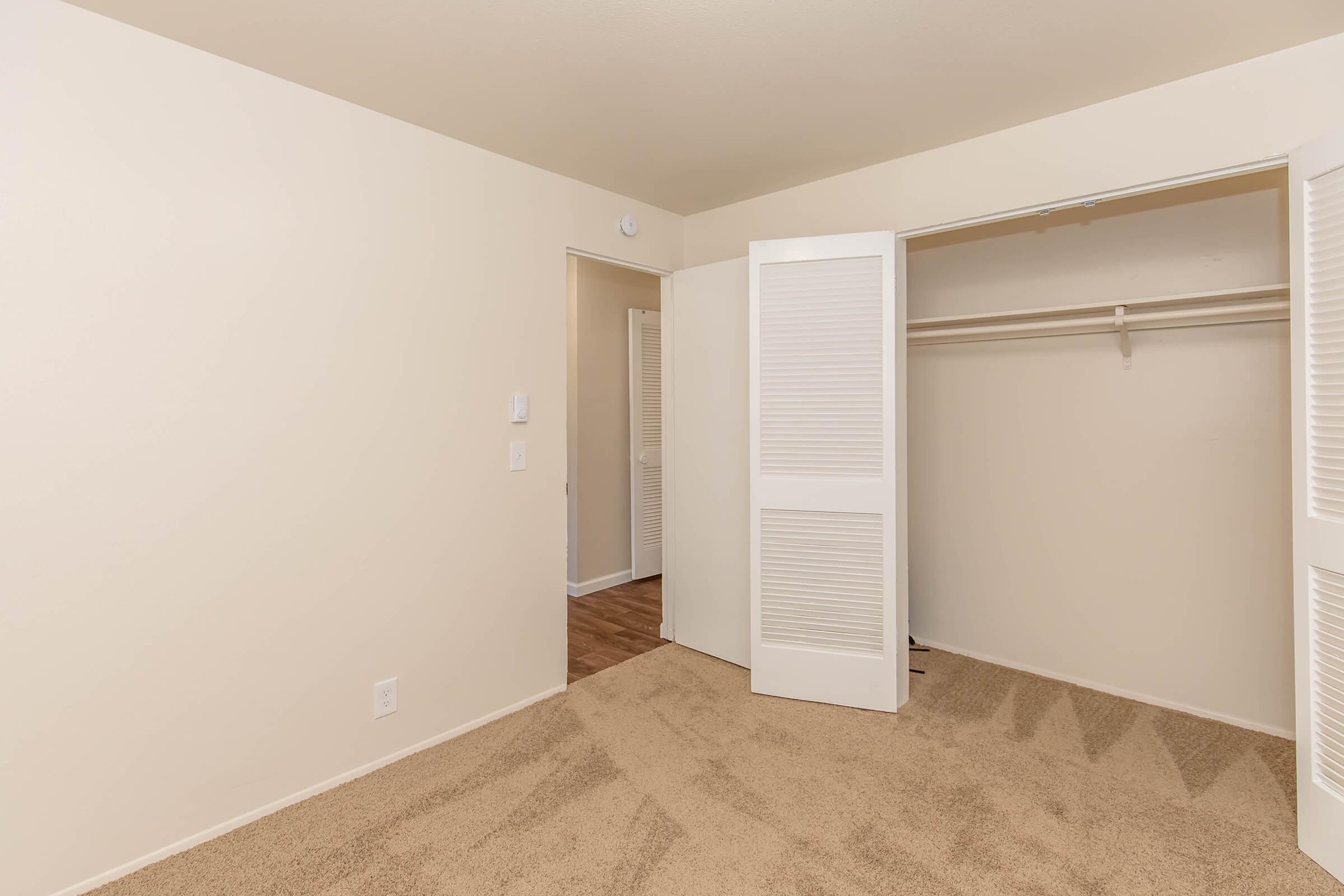 Empty room with tan carpet and beige walls. A closet with white sliding doors is visible, along with a door leading to another space. The room appears well-lit and uncluttered, creating a neutral and spacious atmosphere.