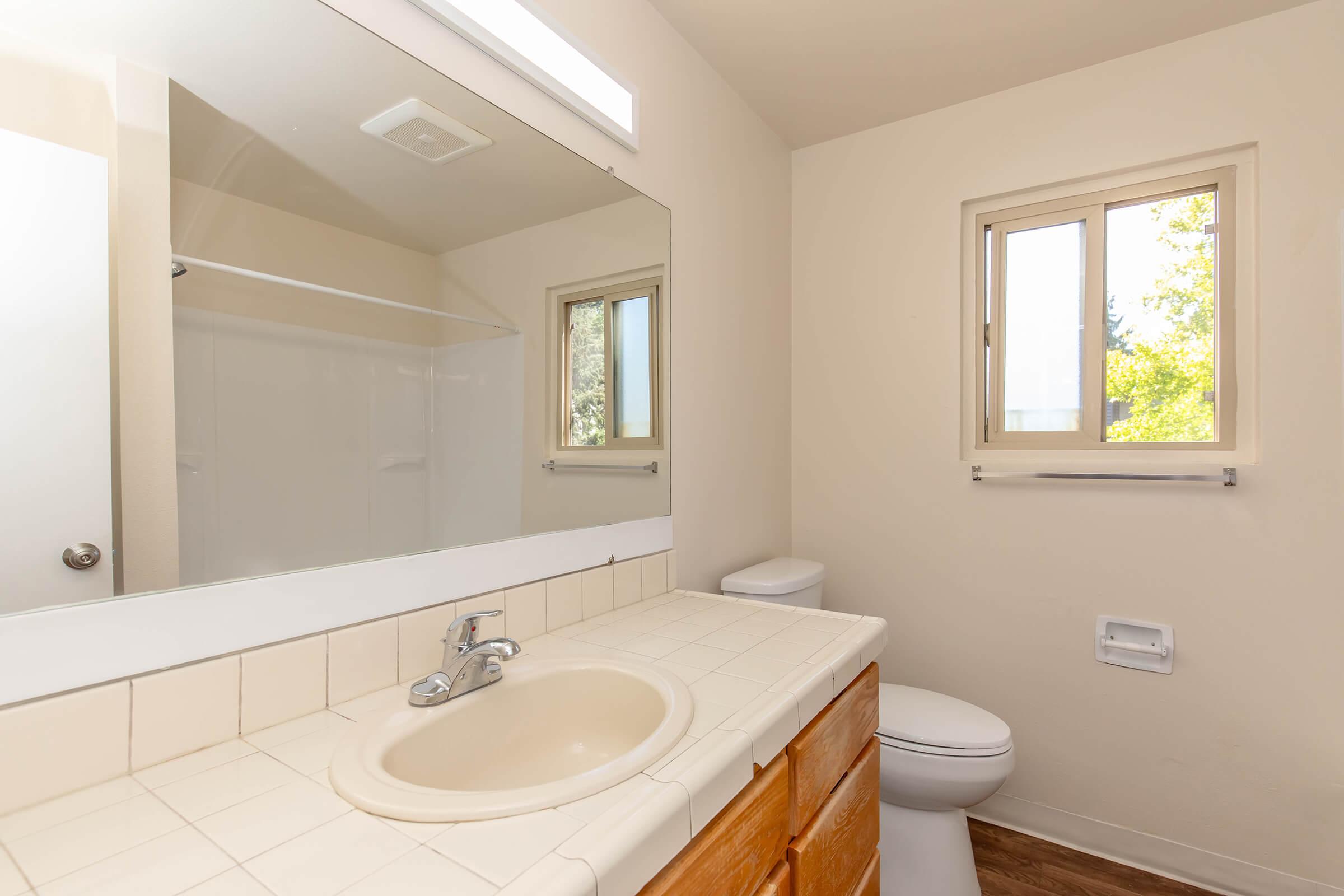 A clean, well-lit bathroom featuring a beige countertop with a sink and faucet, a large mirror above, a window for natural light, and a white toilet. The tiled walls and wooden flooring add a warm touch to the space, with a shower area visible in the background.