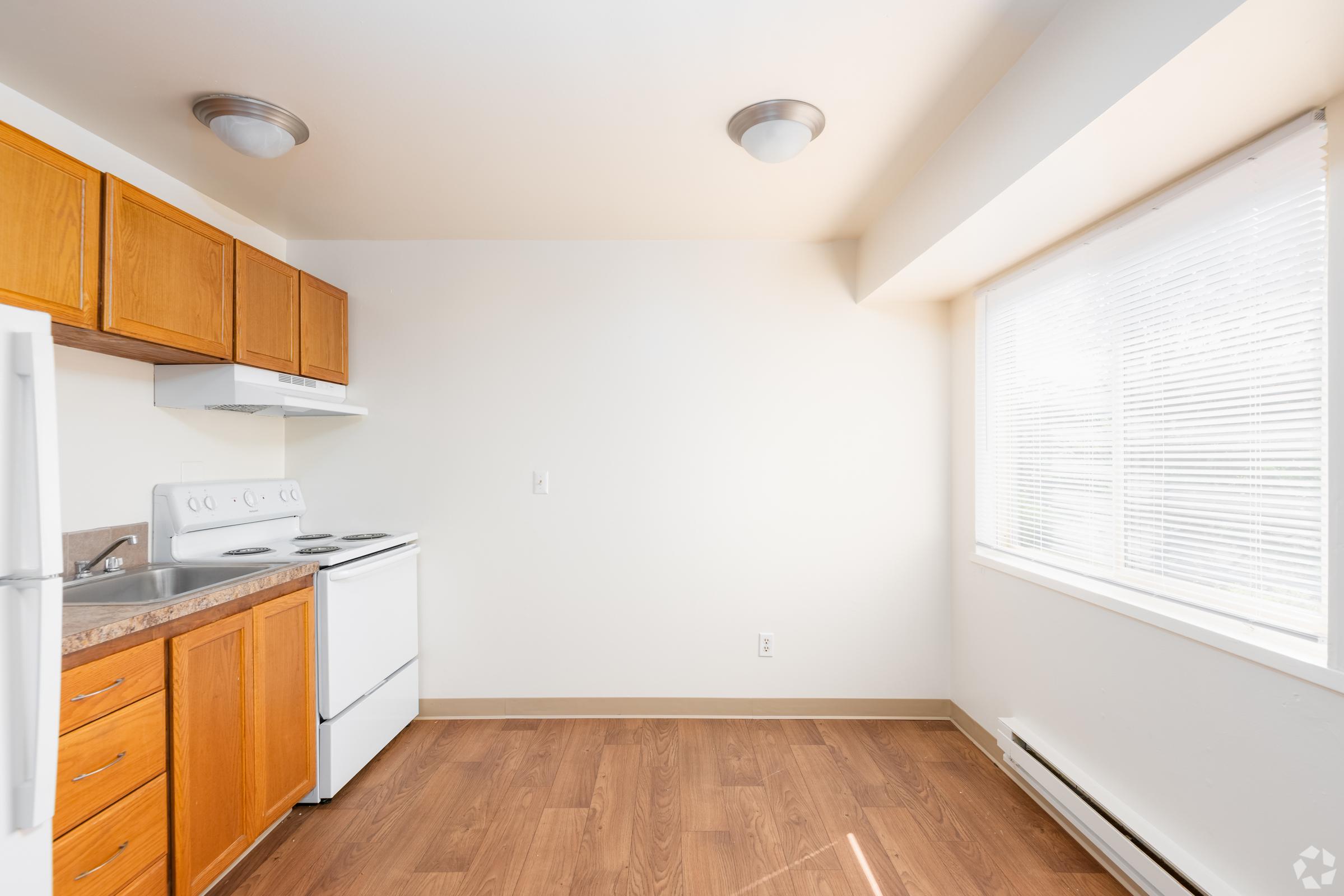 A bright, modern kitchen featuring brown wooden cabinets, a white stove and oven, and a refrigerator. The room has large windows allowing ample natural light, complementing the warm-toned wooden flooring and light-colored walls. The space appears clean and well-organized, ideal for cooking and dining.