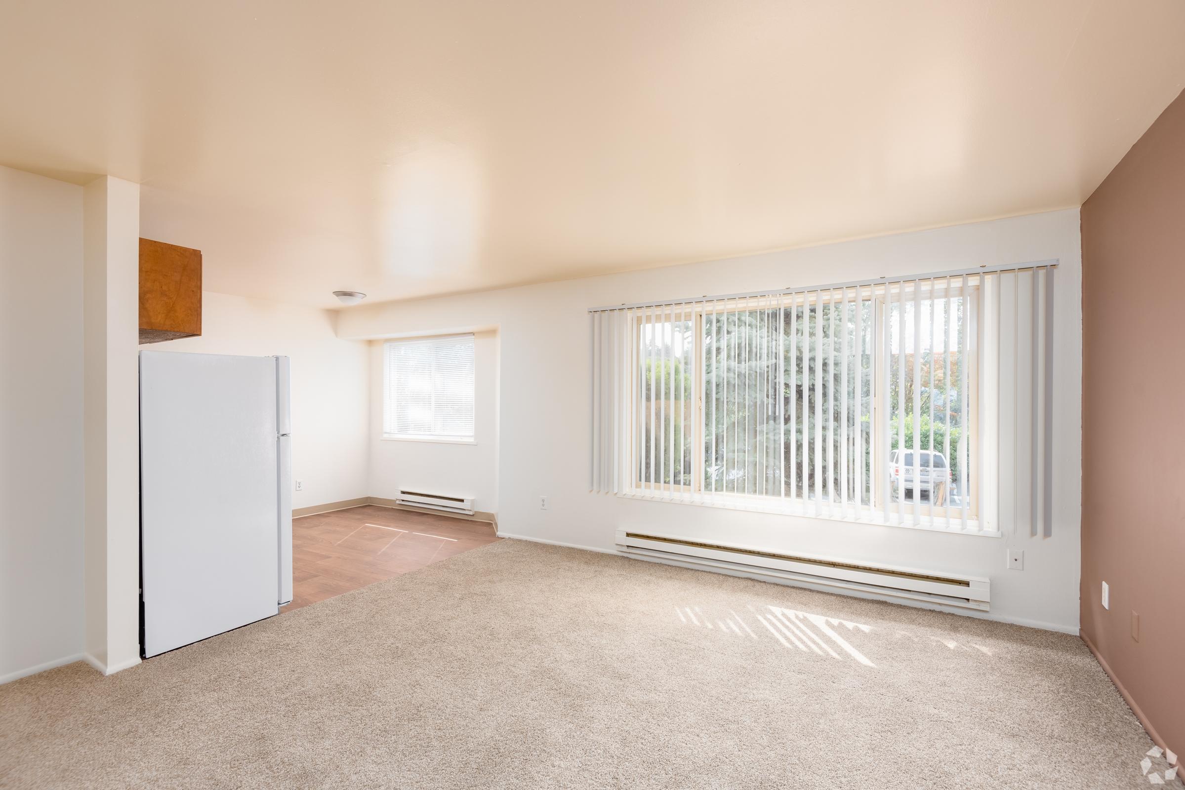 A spacious, empty living area featuring light beige carpet and white walls. A large window with vertical blinds allows natural light to enter, and a white refrigerator is visible on the left. The room is well-lit, with a small section of wall painted in a warm brown hue.