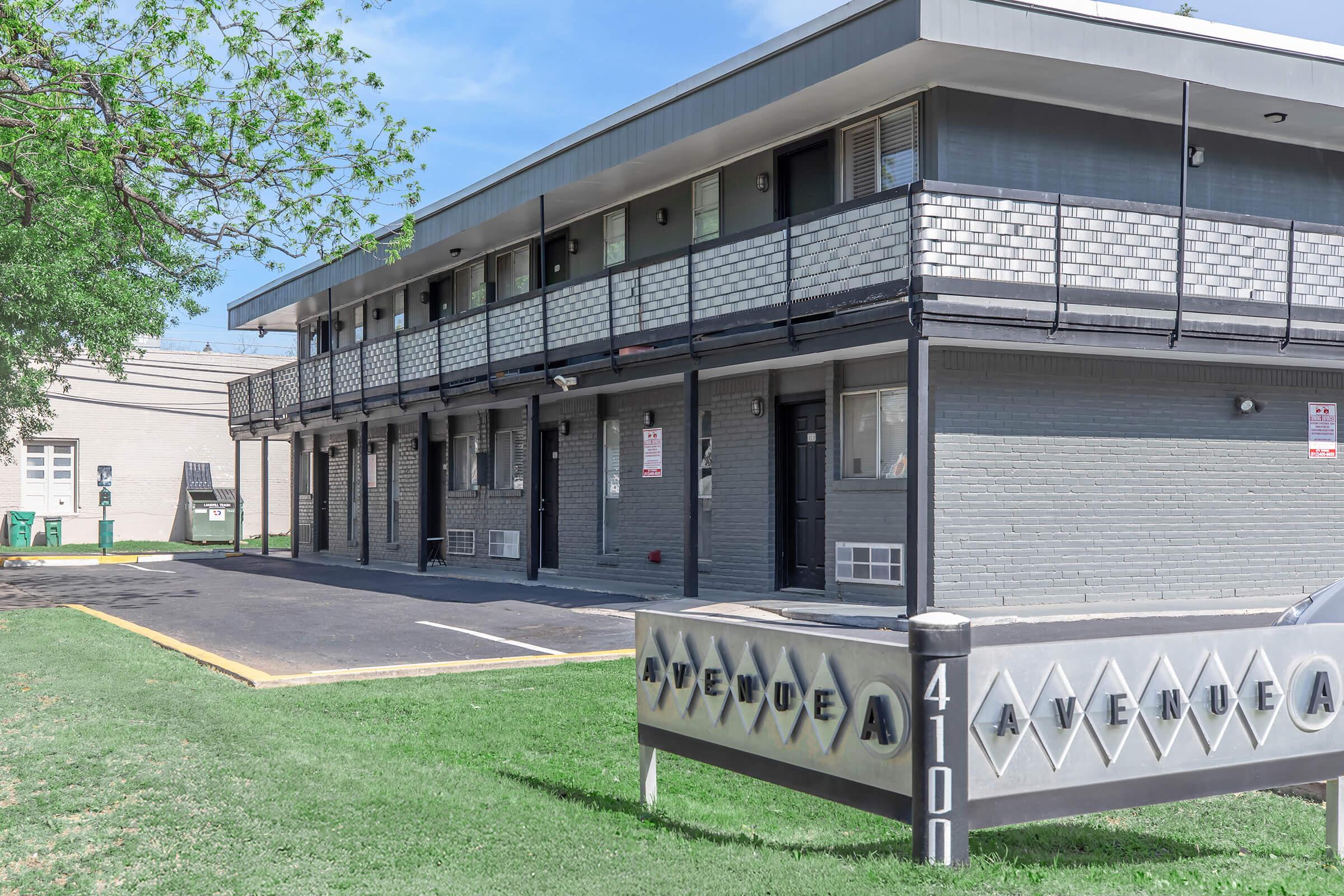 A view of a two-story motel with a gray exterior and black railings. The building has several doors leading to individual rooms, and a sign in front indicating "Avenue A" with the number 4100. There is a grassy area in front and additional structures visible in the background.