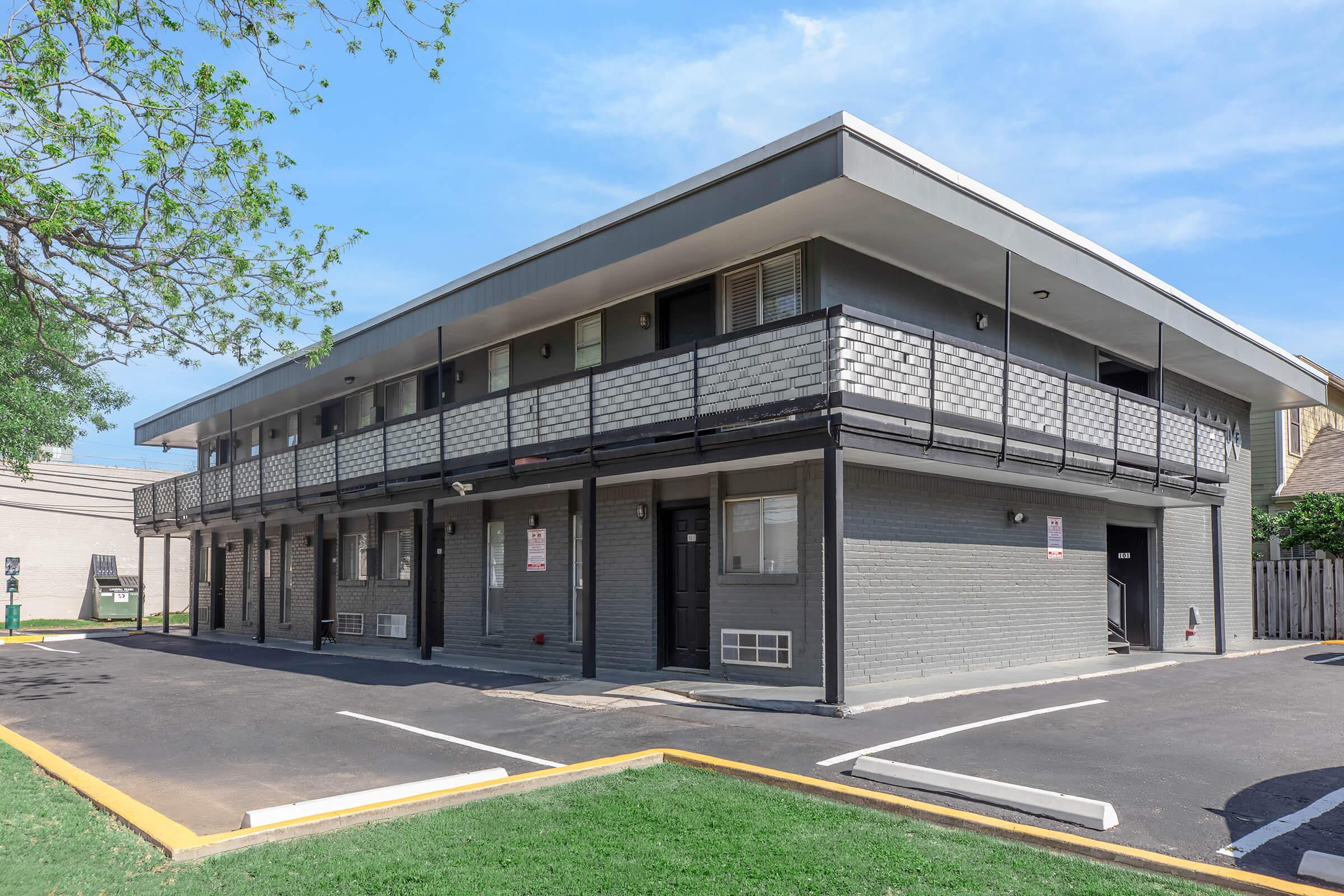A two-story gray motel building with a modern design. It features multiple rooms with balconies, windows, and doors. The surrounding area includes a paved parking lot with marked spaces and a well-maintained green lawn under a clear blue sky.