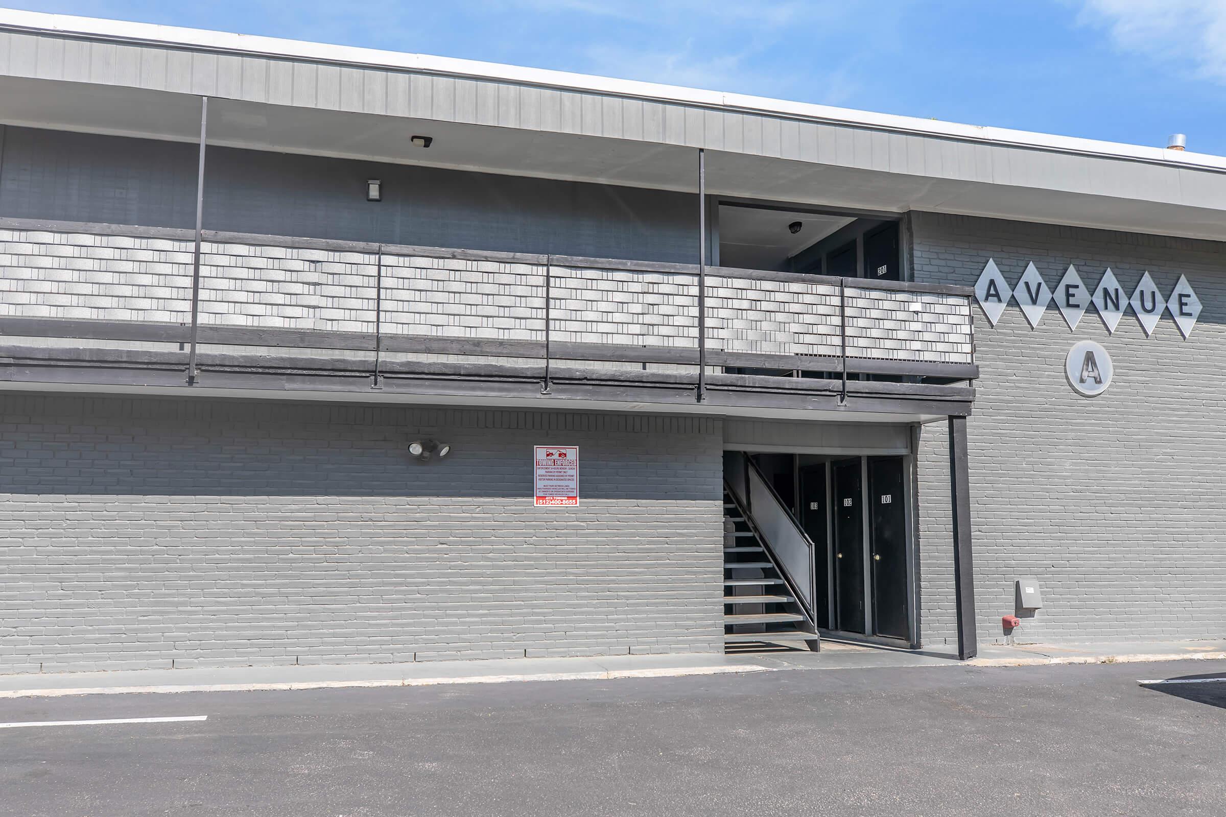 Exterior view of a modern building with a gray façade. The entrance features a staircase and a sign reading "AVENUE." There are windows on the upper level and a sign about regulations posted near the entrance. The sky is clear, adding to the bright atmosphere.