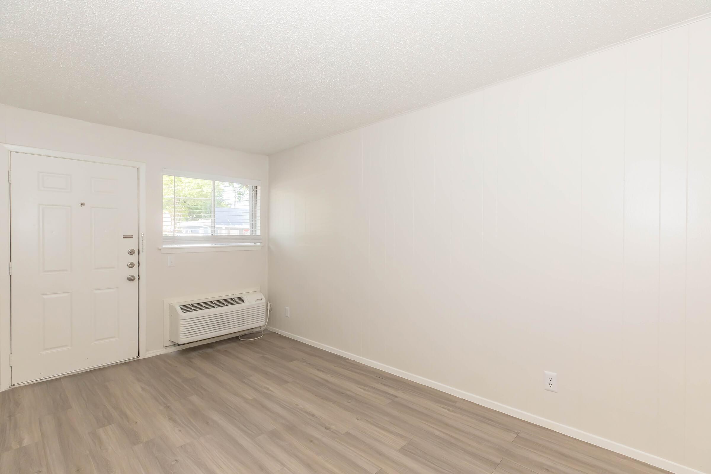 Empty room with light-colored walls and a window on the left side. A front door is visible, and there is a wall-mounted air conditioning unit beneath the window. The floor is covered with light wooden laminate, providing a clean and simple aesthetic.