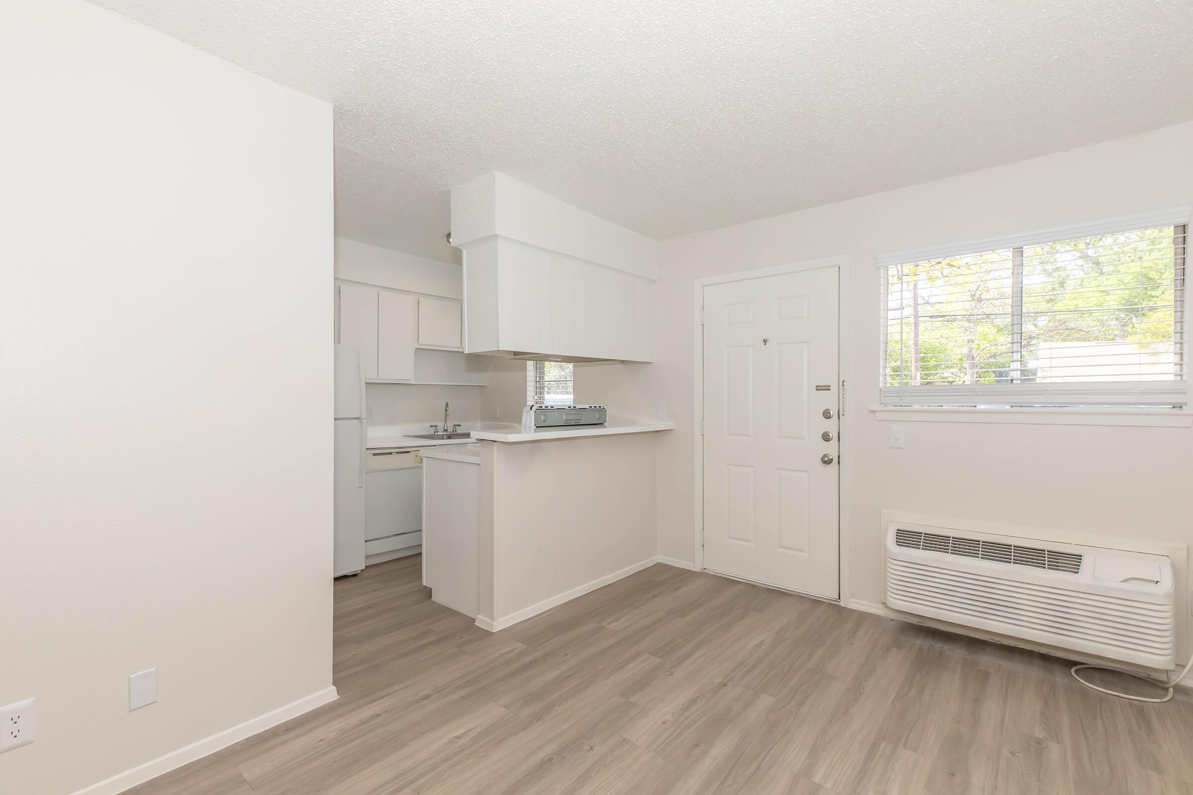 Bright and airy interior of an apartment showcasing a small kitchen space with white appliances, a living area with neutral-colored walls, and large windows that let in natural light. Features include light wood-like flooring and a wall-mounted air conditioning unit.