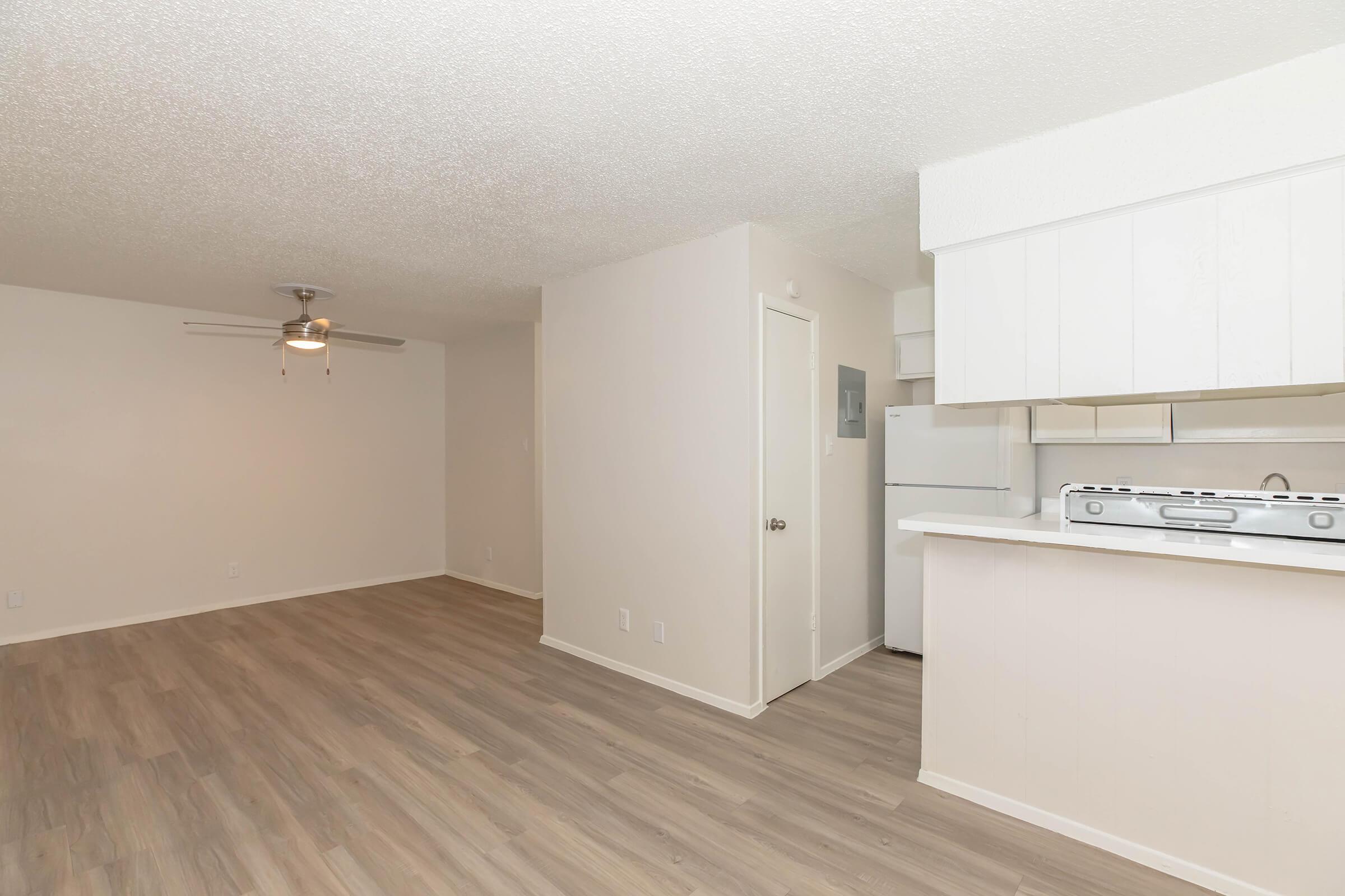 Light and airy living space featuring a ceiling fan, beige walls, and laminate flooring. The open layout includes a view of the kitchen with a refrigerator and gas stove, creating a cozy atmosphere perfect for a small apartment. Natural light fills the room, enhancing the spacious feel.