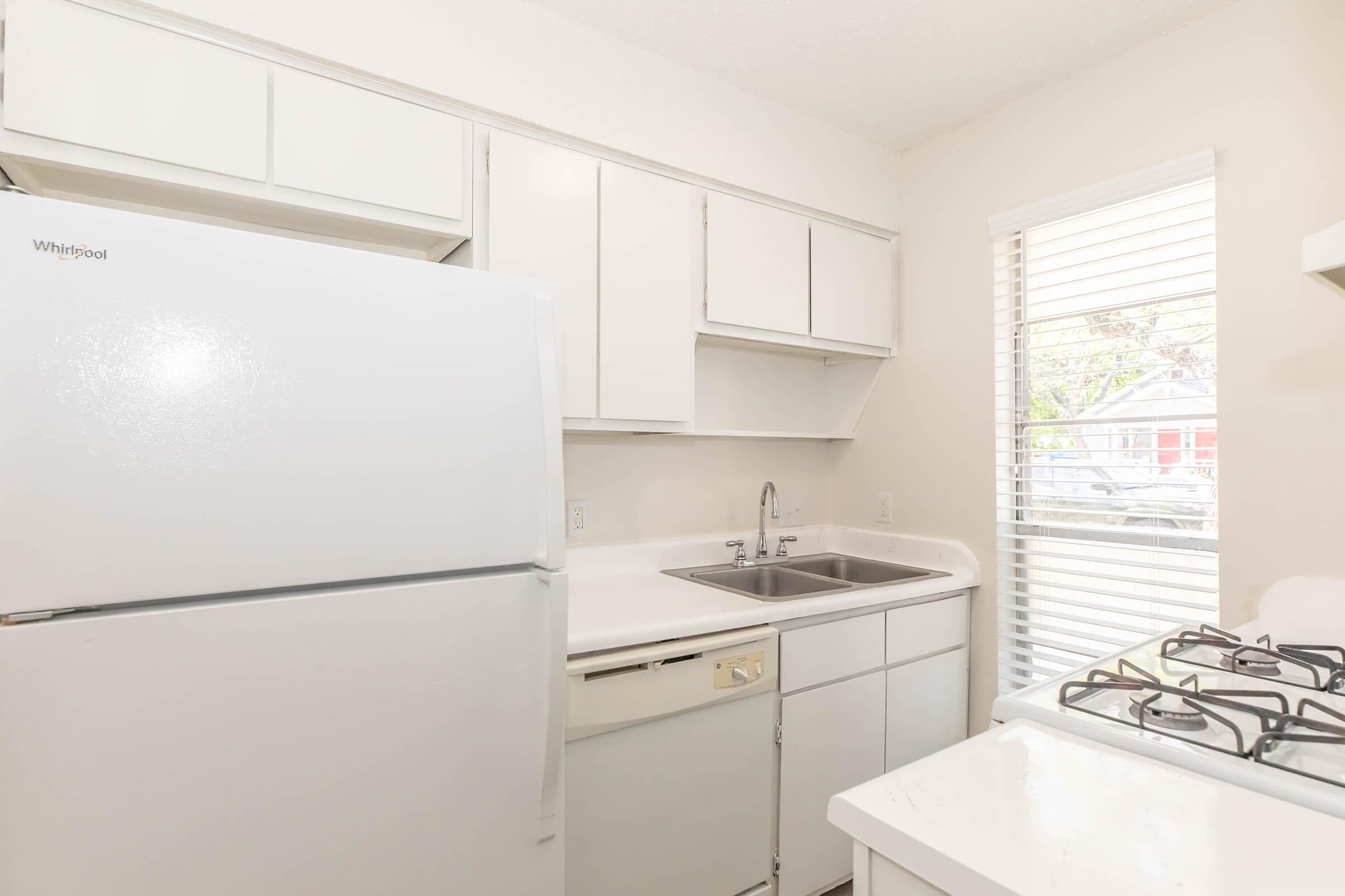 A bright, modern kitchen with white cabinets and appliances. Features a white refrigerator, a sink with a faucet, and a stove with a gas burner. Natural light streams through a window with blinds, illuminating the clean, minimalist design.