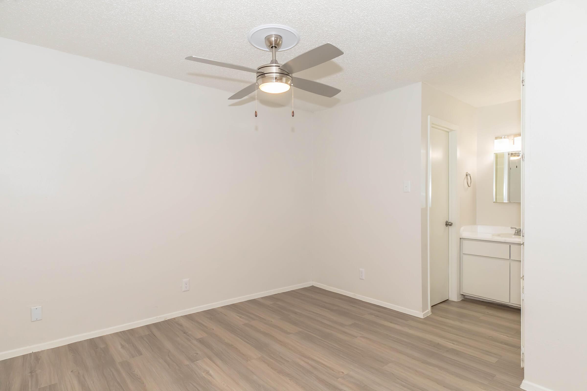 A ceiling fan in a light-colored room with beige walls and brown laminate flooring. The space appears empty with a small bathroom visible in the background, featuring a mirror and sink. Natural light brightens the area, creating a clean and modern atmosphere.