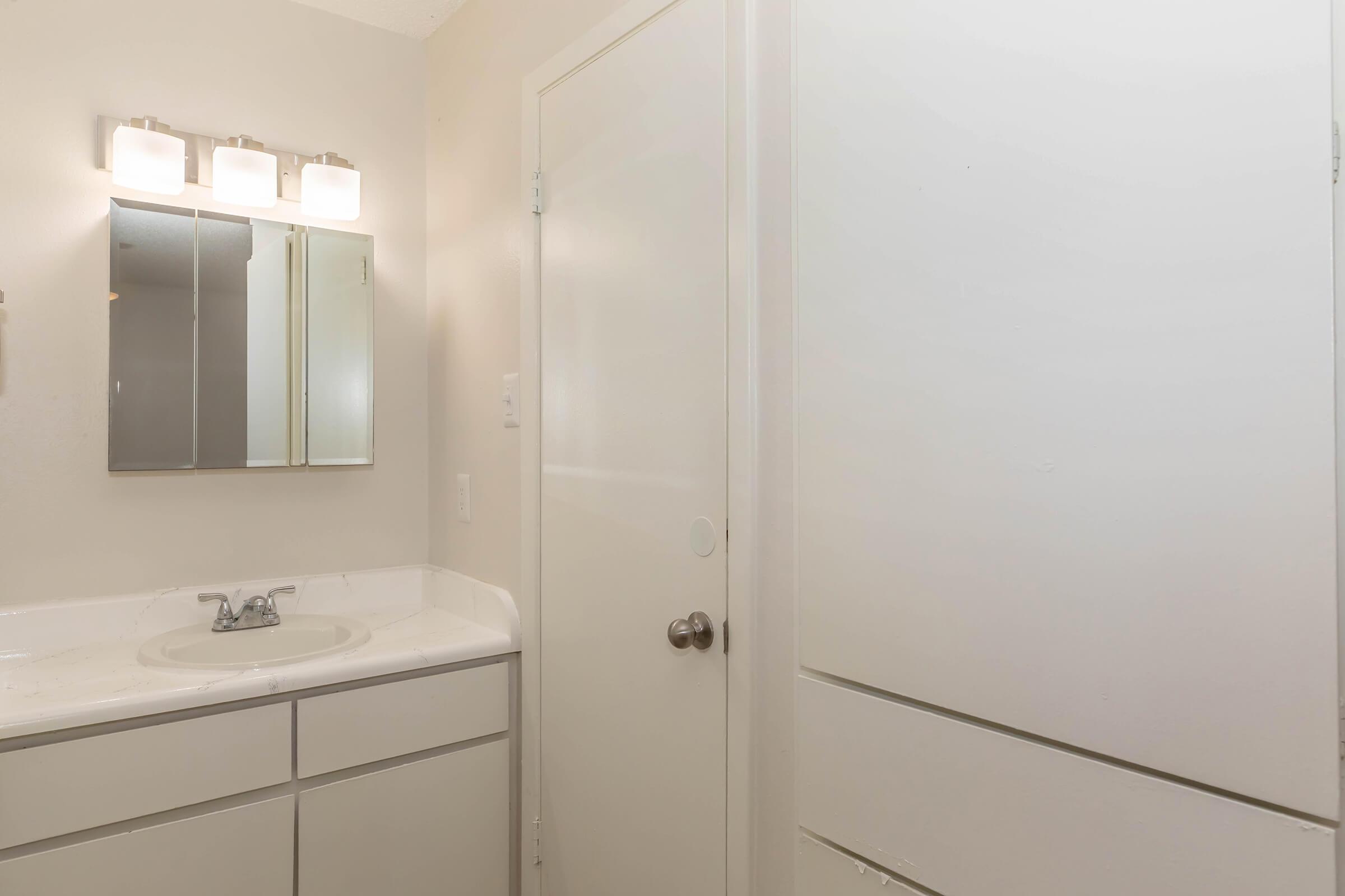 A simple bathroom interior featuring a white vanity with a sink, a mirror above, and a light fixture. There is a closed door leading to another area, and a tall white cabinet beside it. The walls are painted in a light color, creating a clean and minimalistic look.