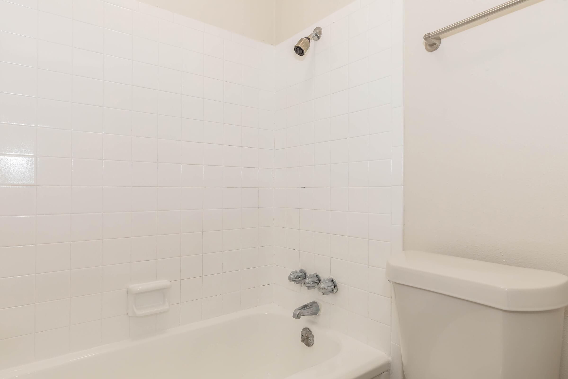 A clean and simple bathroom featuring a white tiled wall, a bathtub with silver faucets, and a toilet. The design is minimalist with bright lighting, contributing to a fresh and spacious appearance.