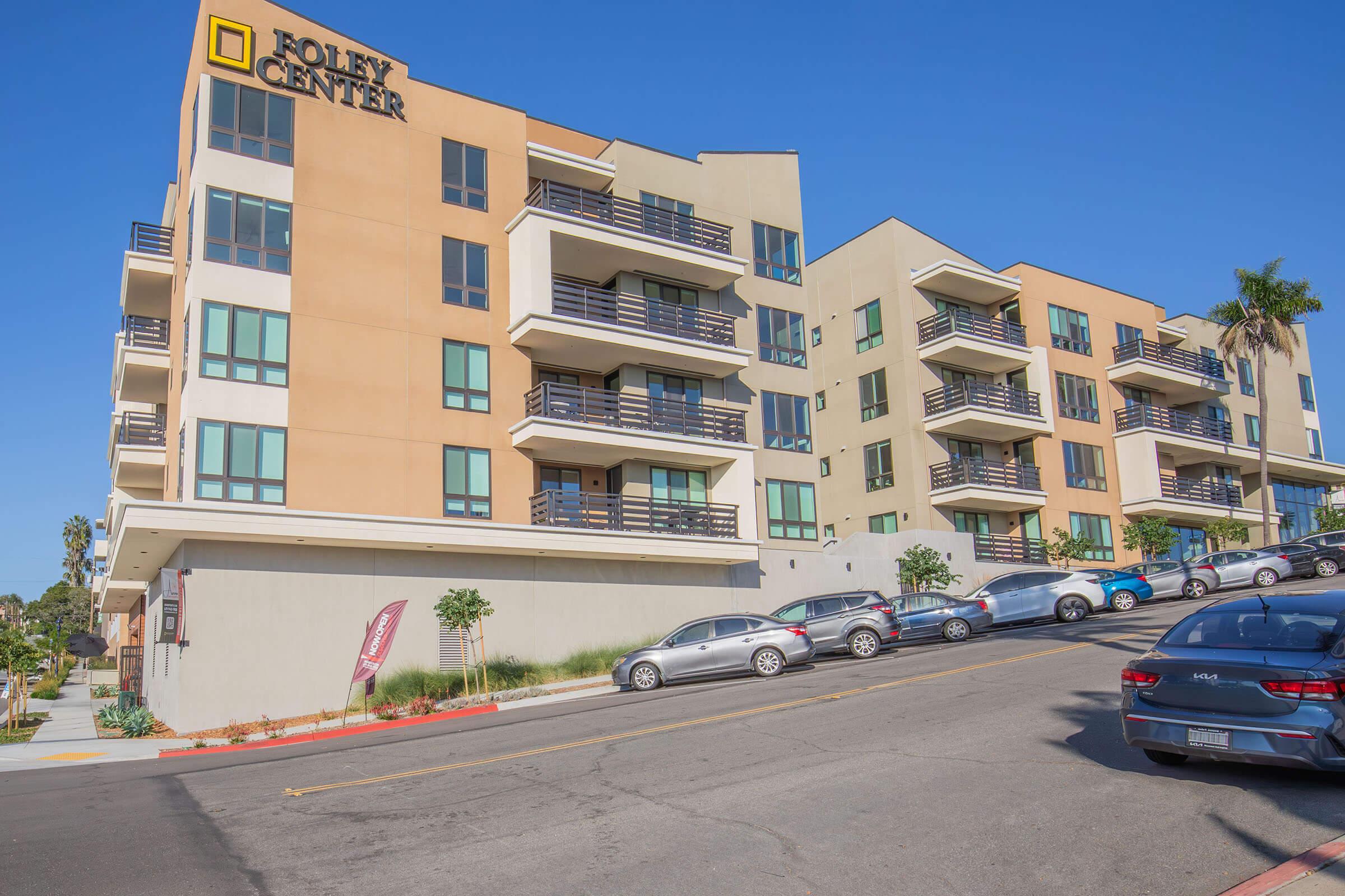 A multi-story building labeled "Foley Center" with modern architecture. The structure features a mix of beige and cream-colored walls, balconies, and a series of parked cars along a sloped street. Clear blue skies provide an inviting backdrop to the scene.